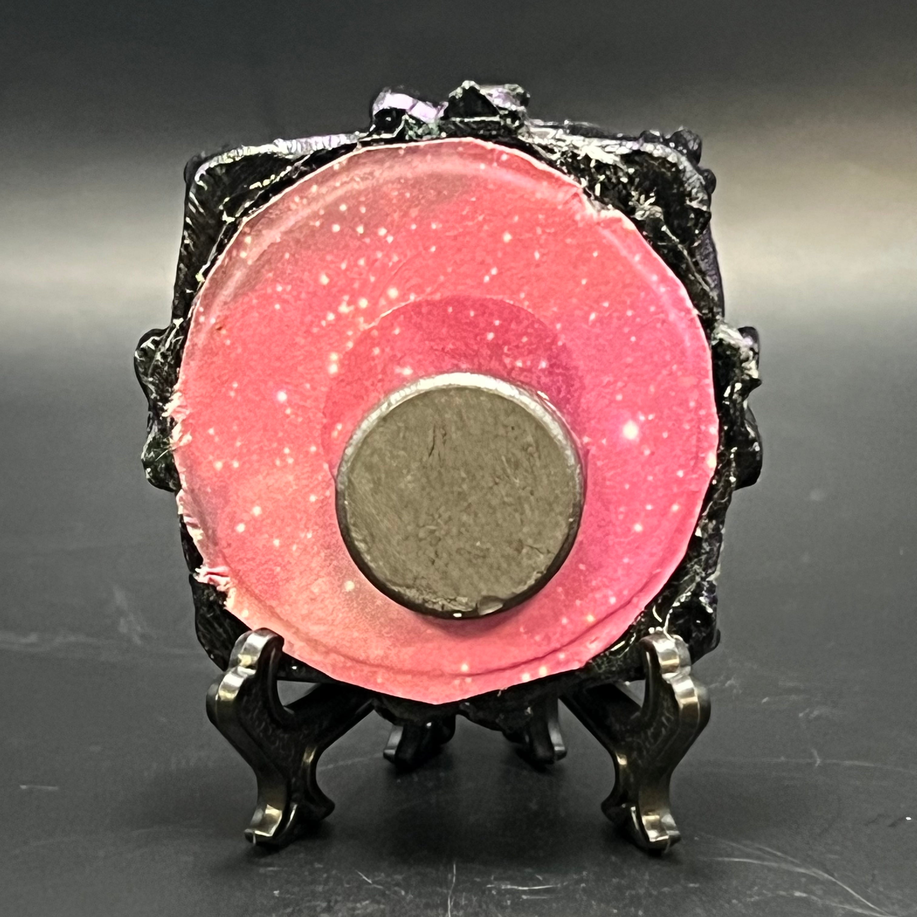 This is the Clustered Crescent Mini Painting by Chris Foster Design. This is showcasing the back. There is a black circular frame ink, on a black stand, a round magnet nearing the middle, backed with purple and hot pink gas nebula origami paper. It is against a dark background.