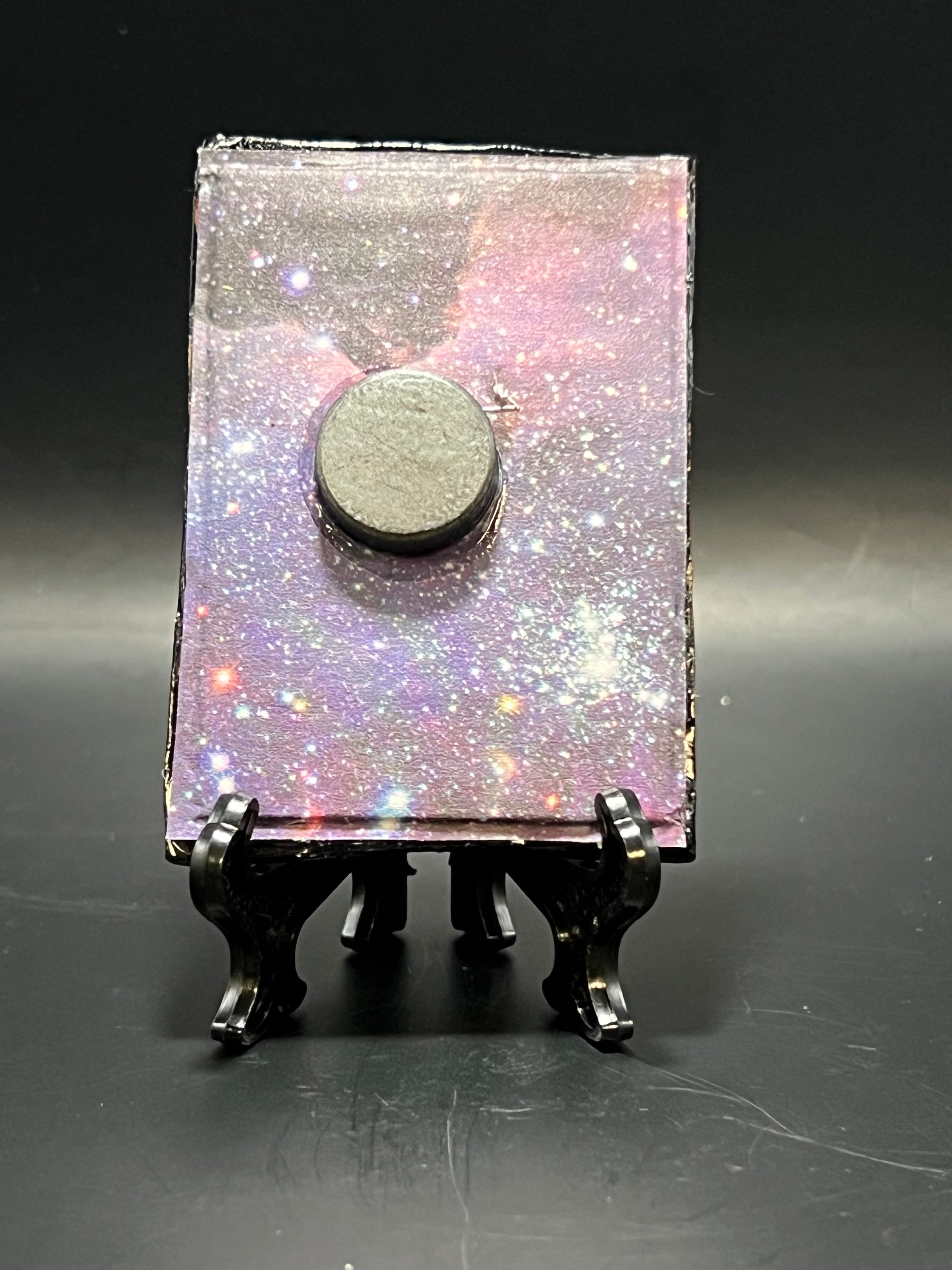 This is the handmade Crescent Moon and Nebula Stars Miniature Painting by Chris Foster Design. This is showcasing the back. This is a small rectangular frame and the decorative item with a sparkly mix background with pink, orange, blue, black and purple on a stand with yellow, red and blue stars. There is a magnet and against a miniature picture frame.