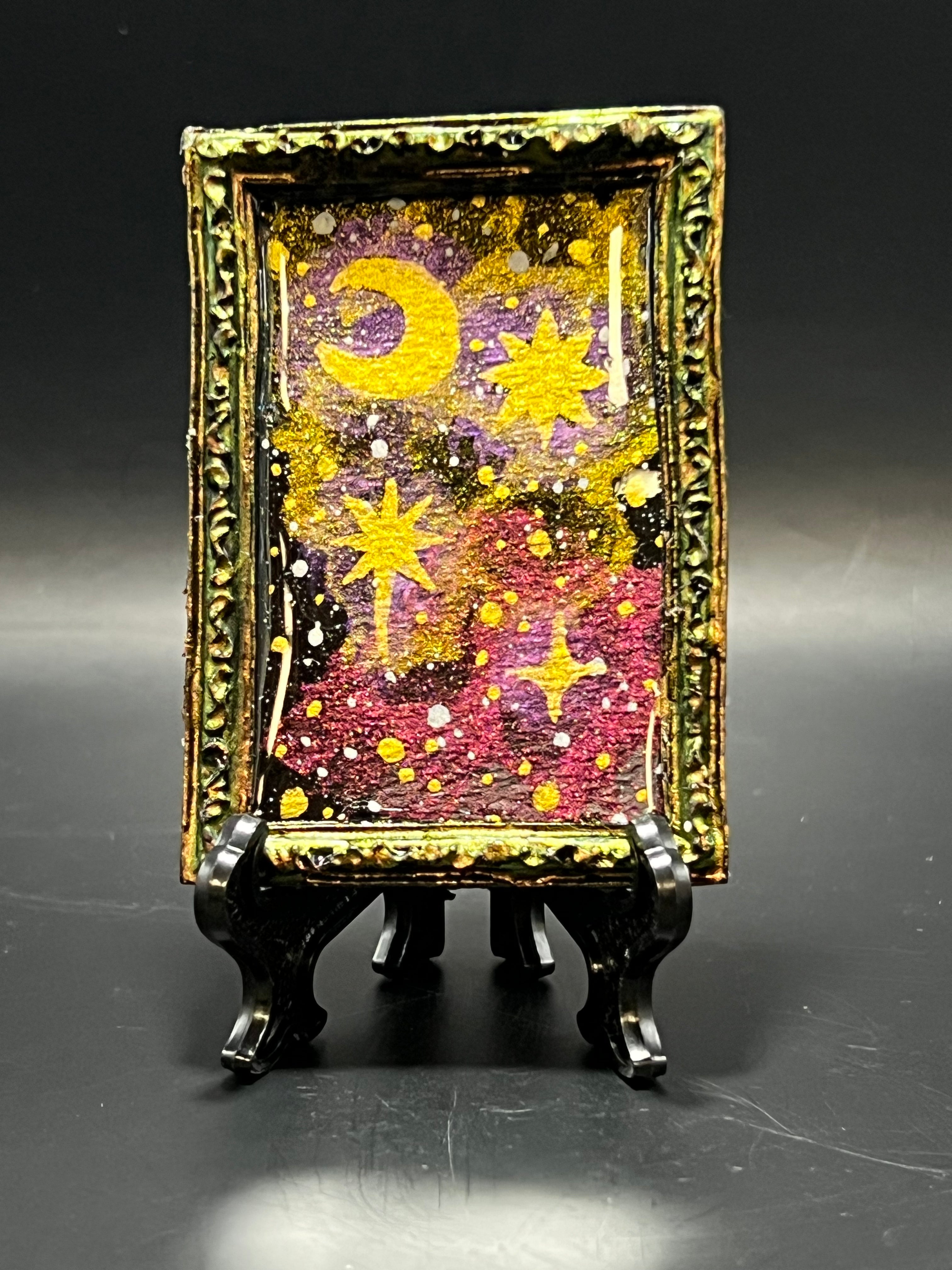 This is the handmade Crescent Moon and Stars Nebula Miniature Painting by Chris Foster Design. This is showcasing the front. This is a decorative piece with a gold rectangular frame, starry night design  with crescent moon to the left including large yellow stars, sparkling  orange, yellow, red and white smaller stars on a dark background. There is a magnet and against a miniature picture frame.