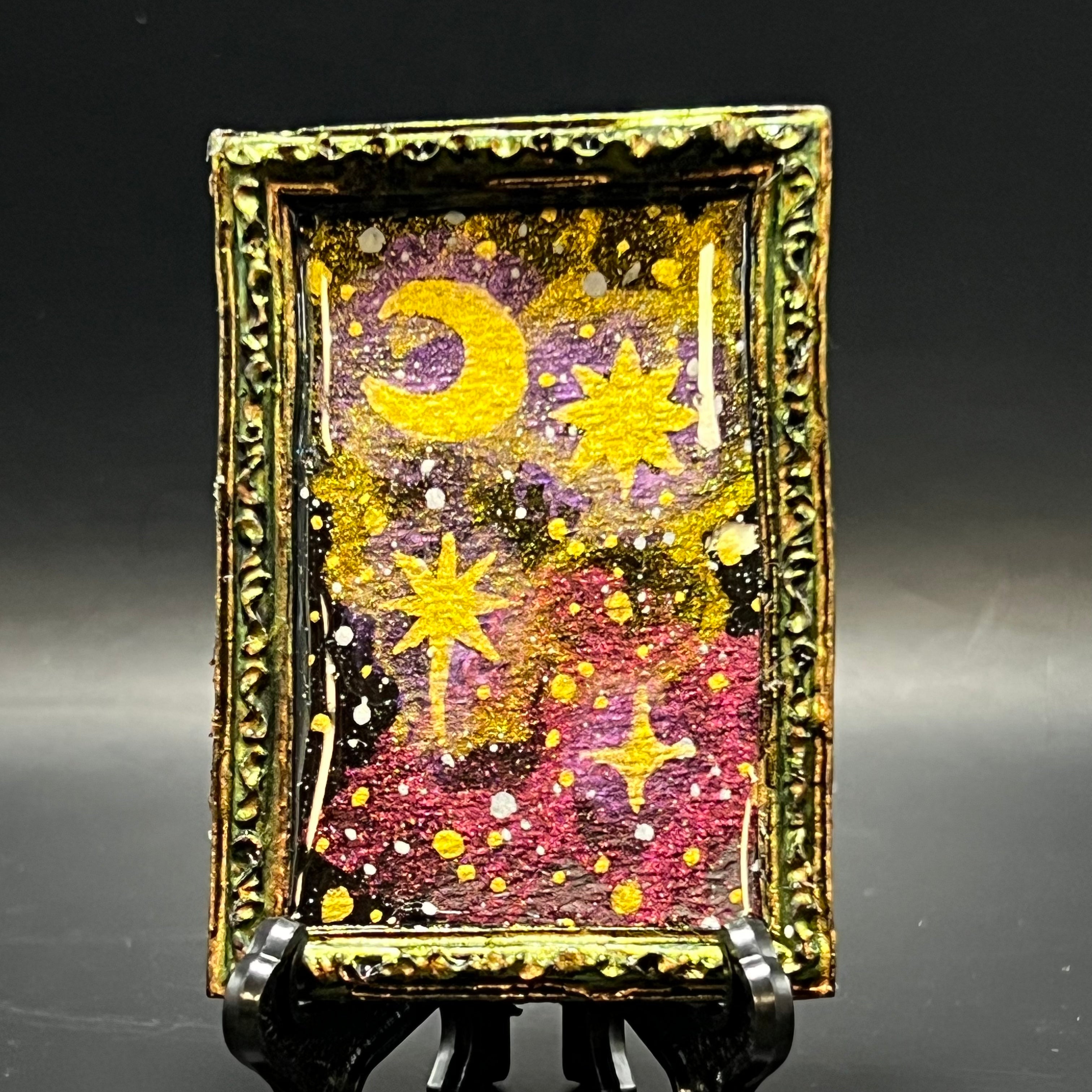This is the Crescent Moon and Stars Nebula Mini Painting by Chris Foster Design. This is showcasing the front. Gold green color shift on black rectangular ornamental frame. Gold green and red purple color shift watercolor nebulae on backdrop of a starry deep space void with glowing golden crescent moon and stars. It is against a dark background and a black stand.