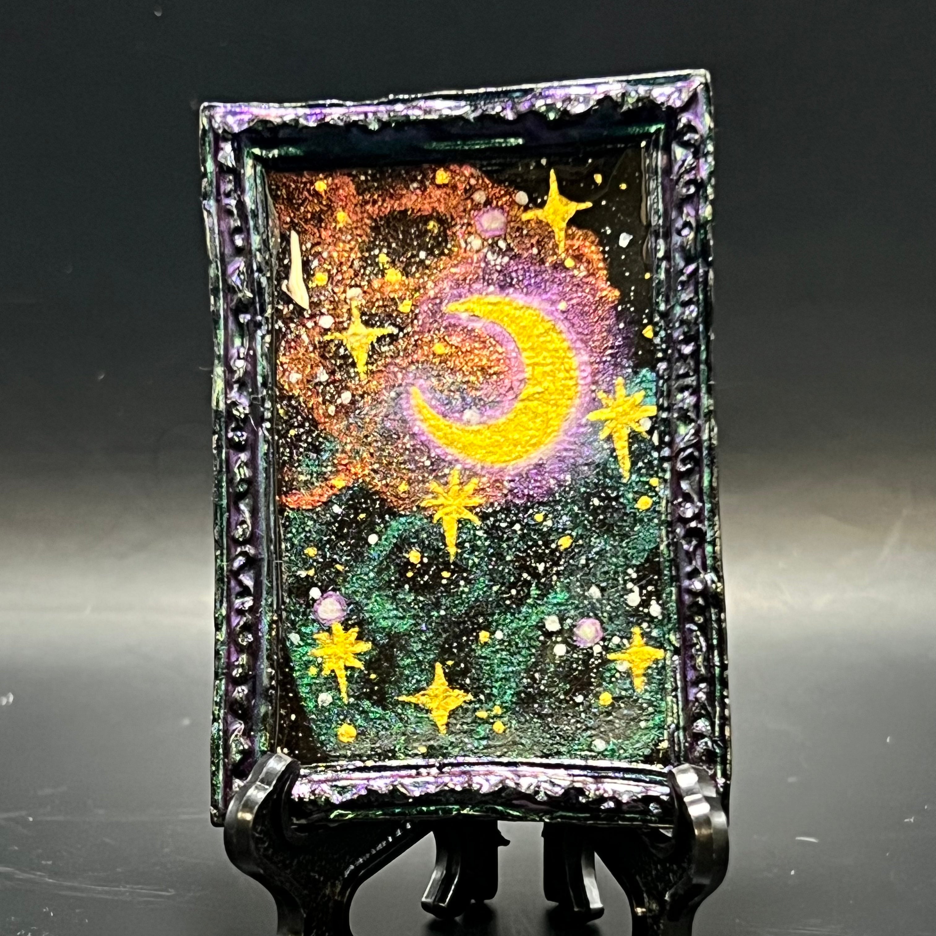 This is the Crescent Nebula Star Burst Mini Painting by Chris Foster Design. This is showcasing the front. Red purple color shift coated black rectangle frame. Red gold and red purple blue color shift nebulas collide on a starry void. Glowing cold stars suspended in space surround a bright crescent moon. It is against a dark background.