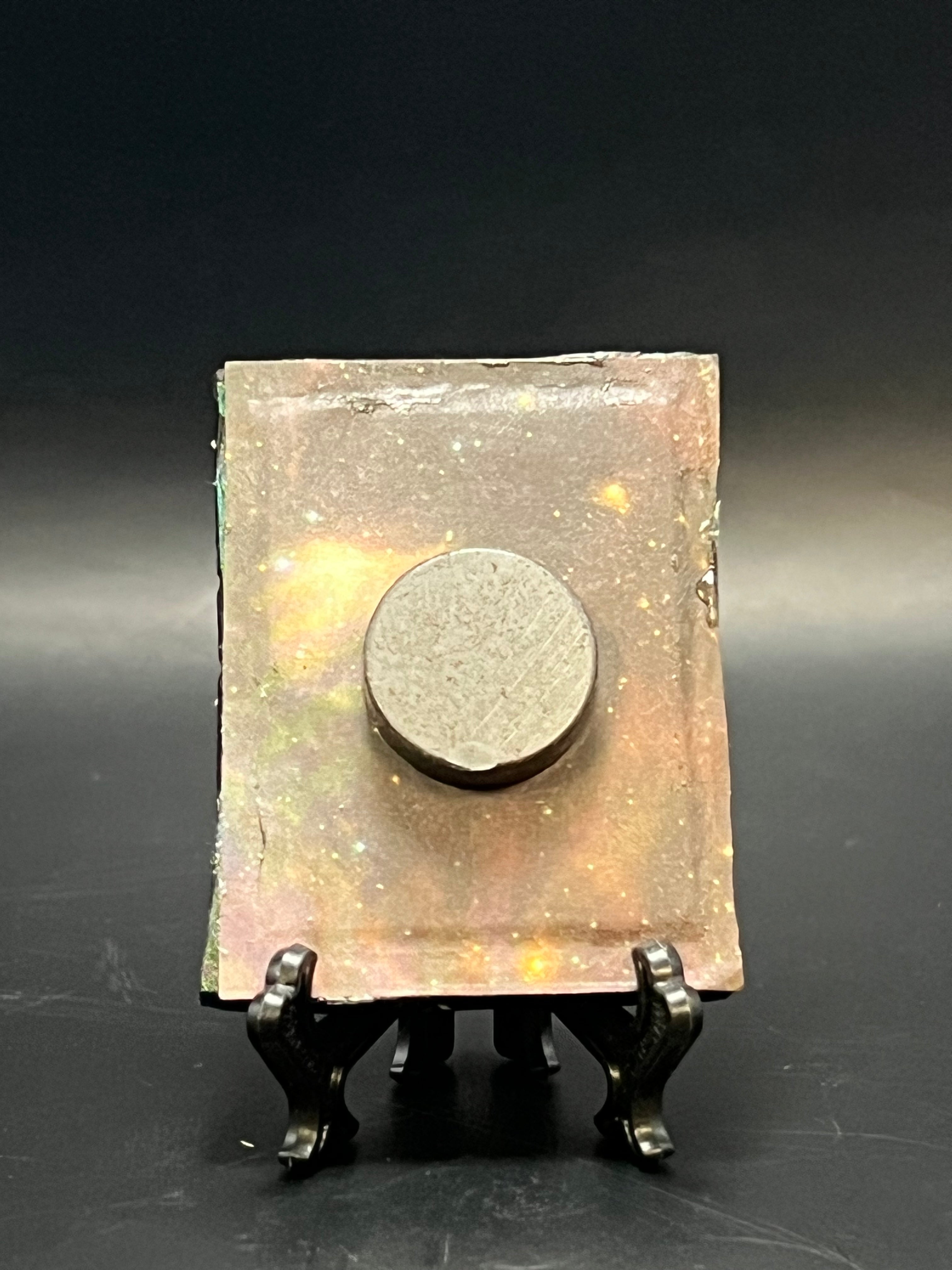 This is the handmade Distant Ringed Planet Miniature Painting by Chris Foster Design. This is showcasing the back.  This is a small rectangular frame and the decorative item with a sparkly mix background with orange, yellow and black on a stand with yellow stars. There is a magnet and against a miniature picture frame.