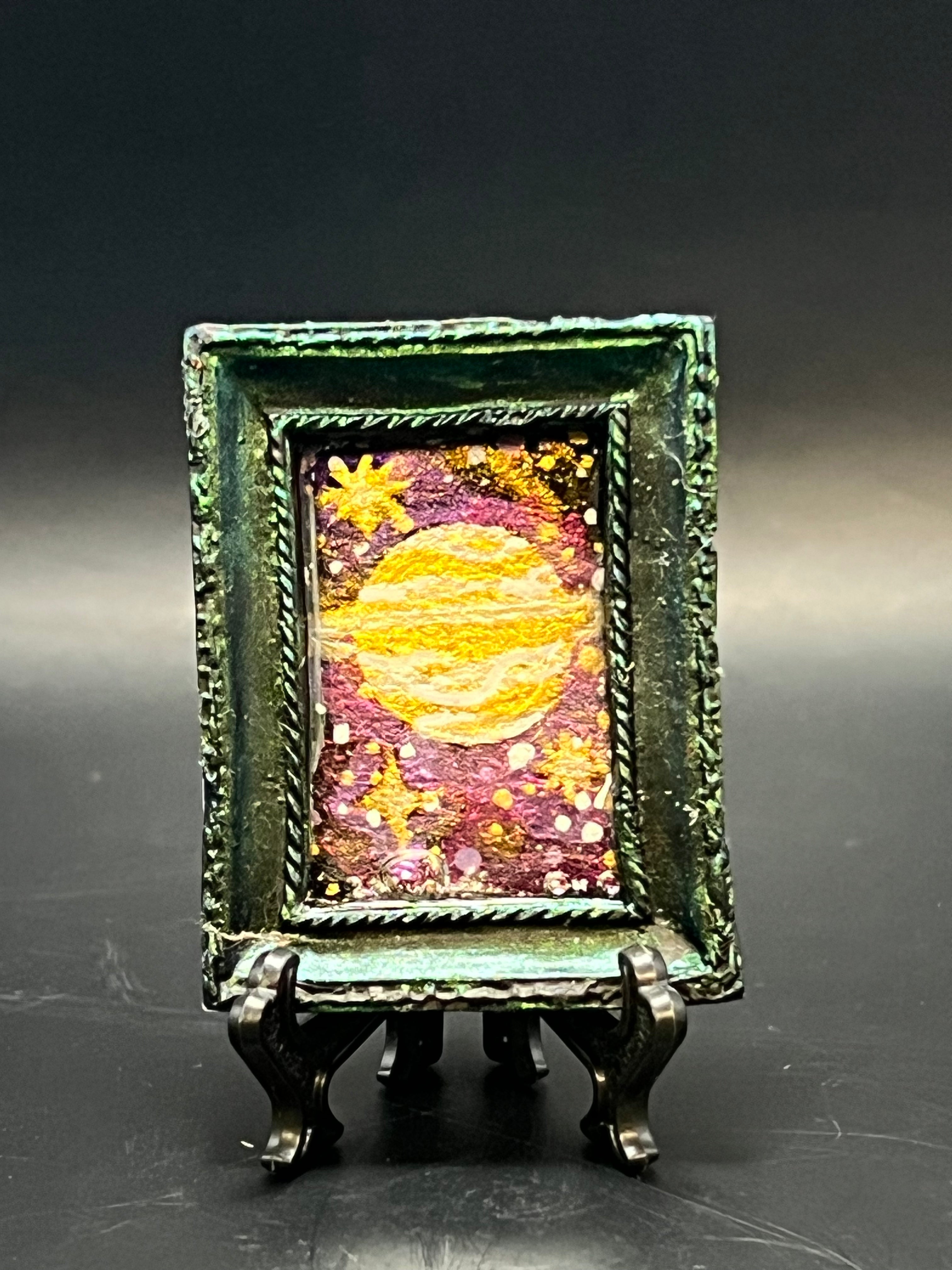 This is the handmade Distant Ringed Planet Miniature Painting by Chris Foster Design. This is showcasing the front. This is a decorative piece with a shimmery green rectangular frame, starry night design including yellow stars, sparkling red, orange, yellow and white stars on a dark background. There is a magnet and against a miniature picture frame.