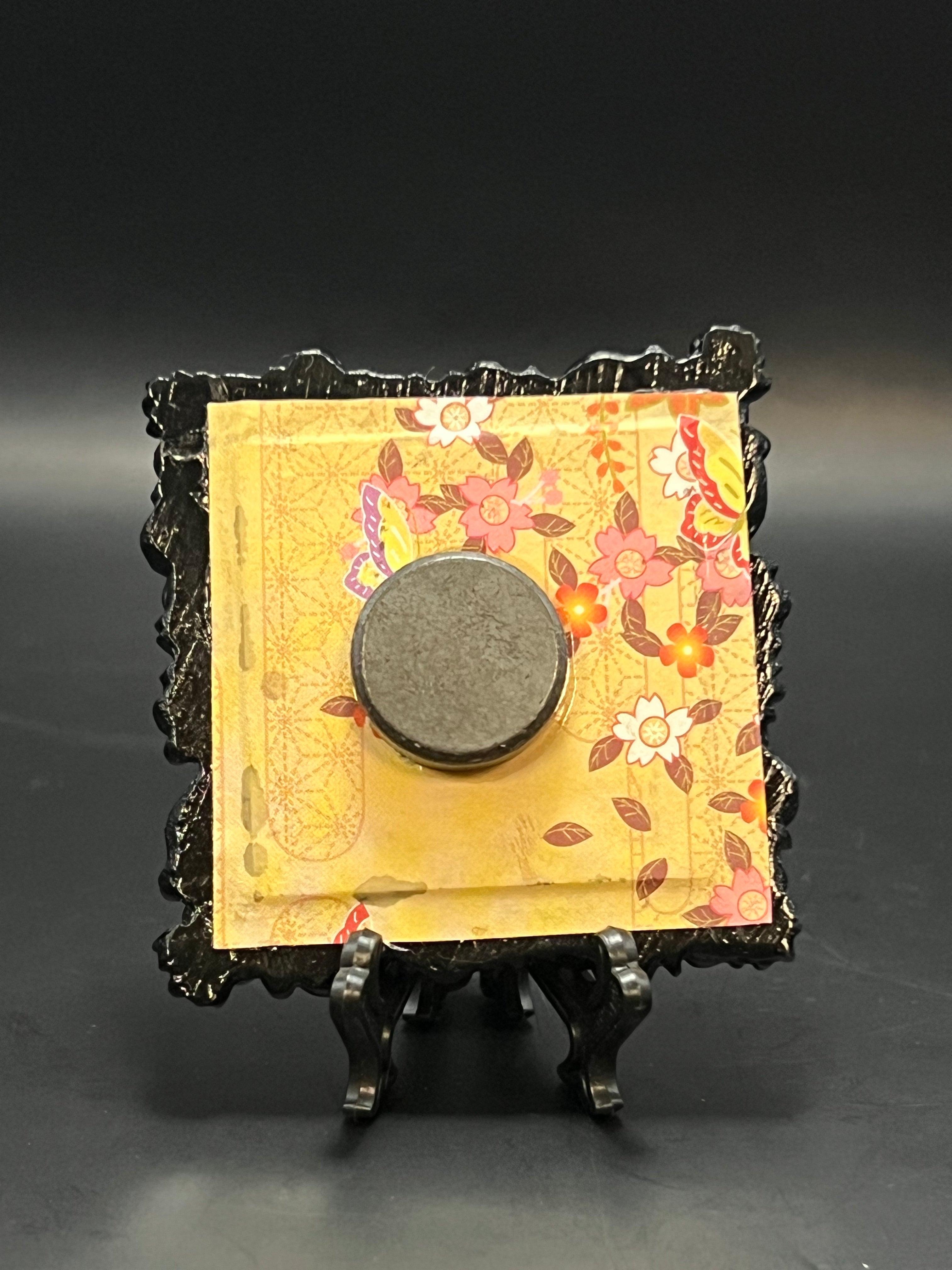 This is the handmade Fly Agaric Mushroom Blue Miniature Painting by Chris Foster Design. This is showcasing the back. This is a small square frame and the decorative item with flowers falling from the right on a yellow background. They are red, white, yellow and maroon flowers with the leaves falling. There is a magnet and against a miniature picture frame.