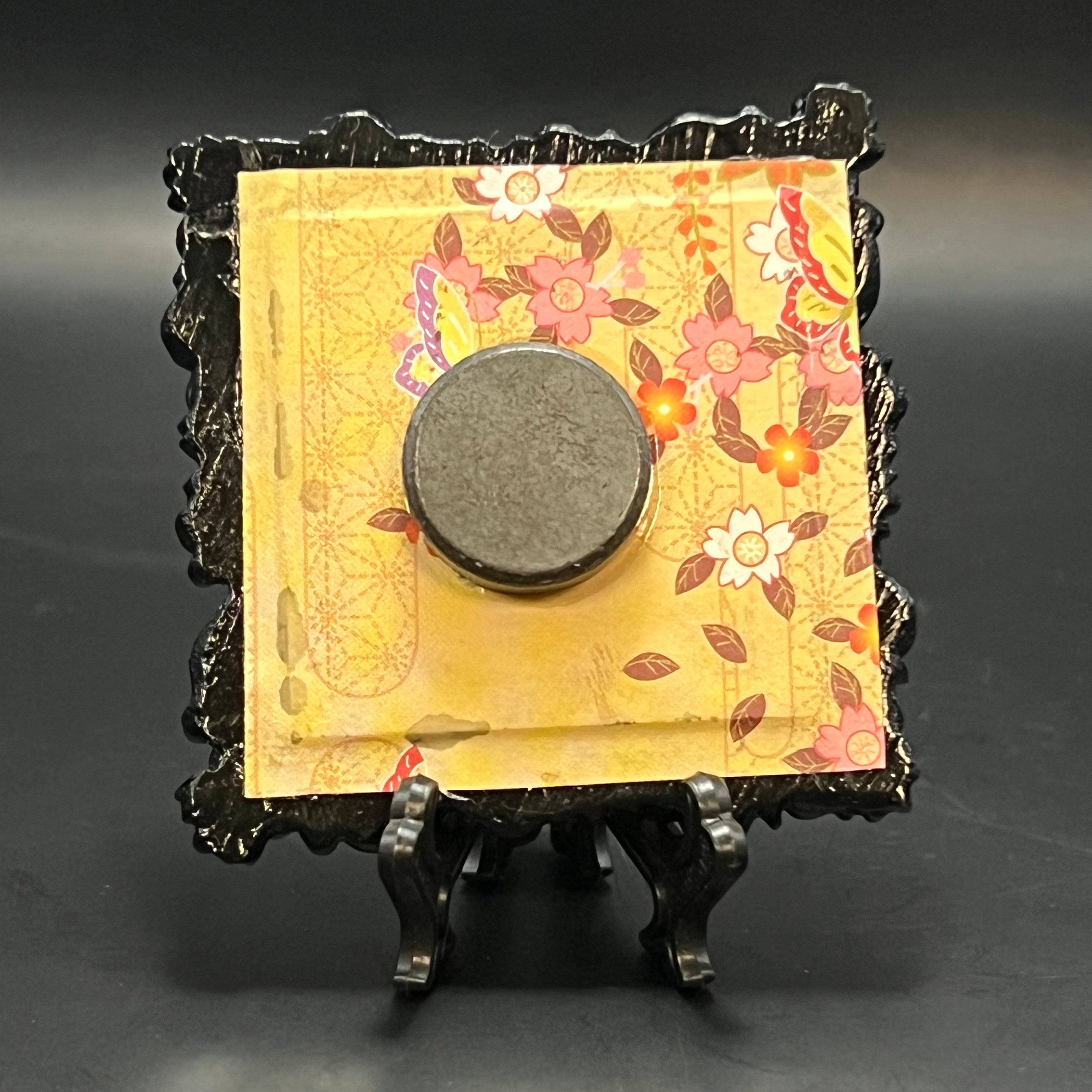 This is the Fly Agaric Mushroom Blue Mini Painting by Chris Foster Design. This is showcasing the back. It is backed with Floral patterned origami paper with a mix of light yellow, orange, pink, purple, brown, and white. There is a round magnet and is on a black stand. It is against a dark background.