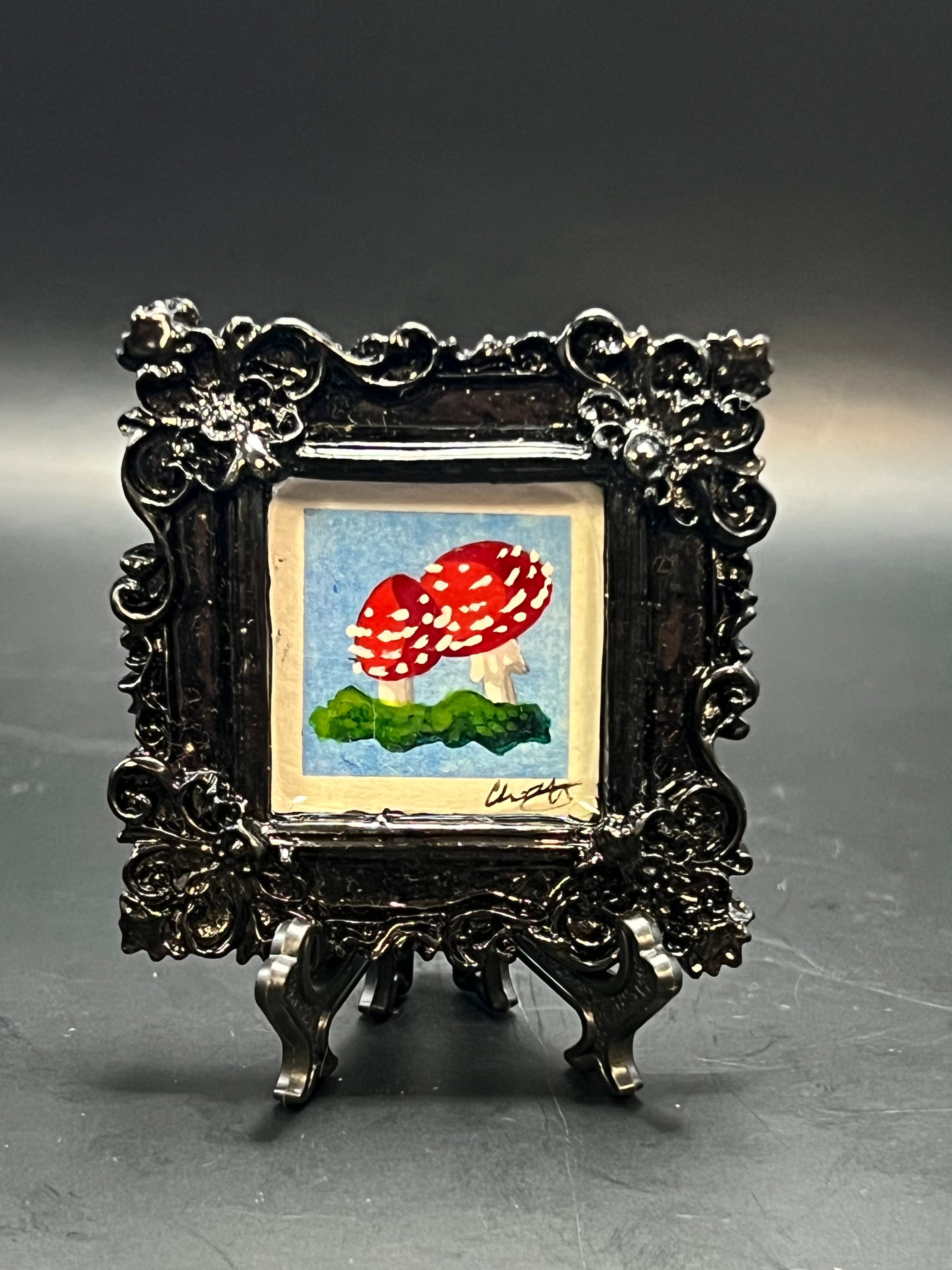 This is the handmade Fly Agaric Mushroom Blue Miniature Painting by Chris Foster Design. This is showcasing the front. This is a decorative piece with a black square frame, there are two mushrooms that are red speckled white on green wavy grass. The painting is against a light blue blackground and signed by the studio. There is a stand that the frame is on.