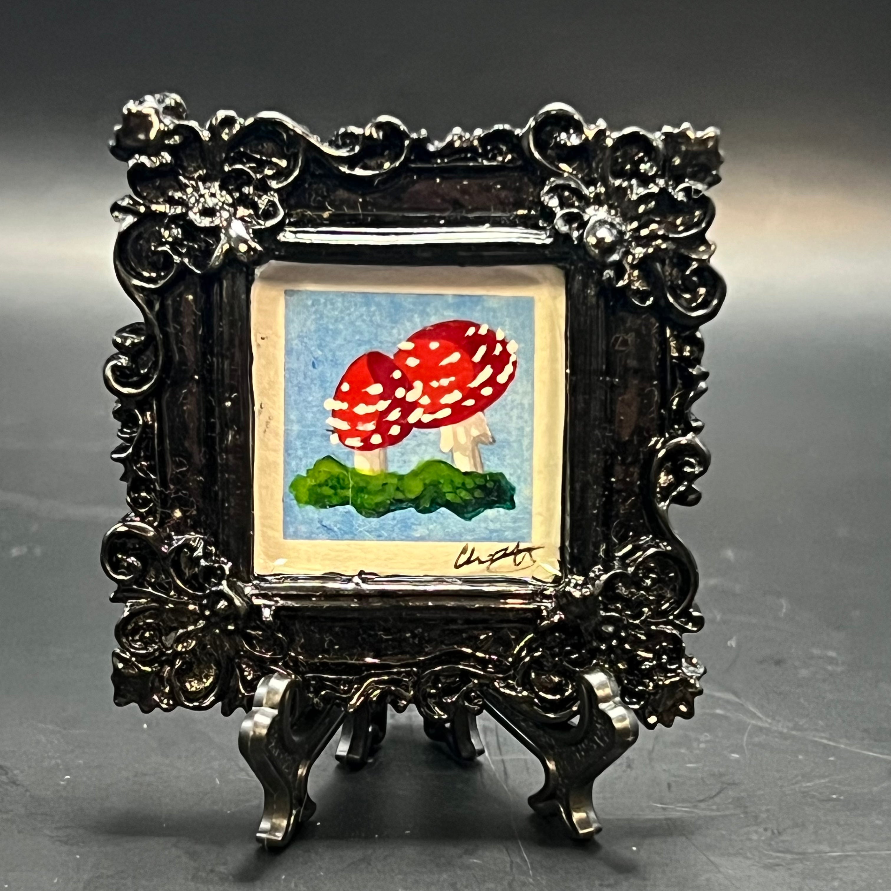 This is the Fly Agaric Mushroom Blue Mini Painting by Chris Foster Design. This is showcasing the front of the painting. Gloss black ornamental square frame. Two red fly agaric mushrooms sprouting from dark and light green moss. It is against a dark background.
