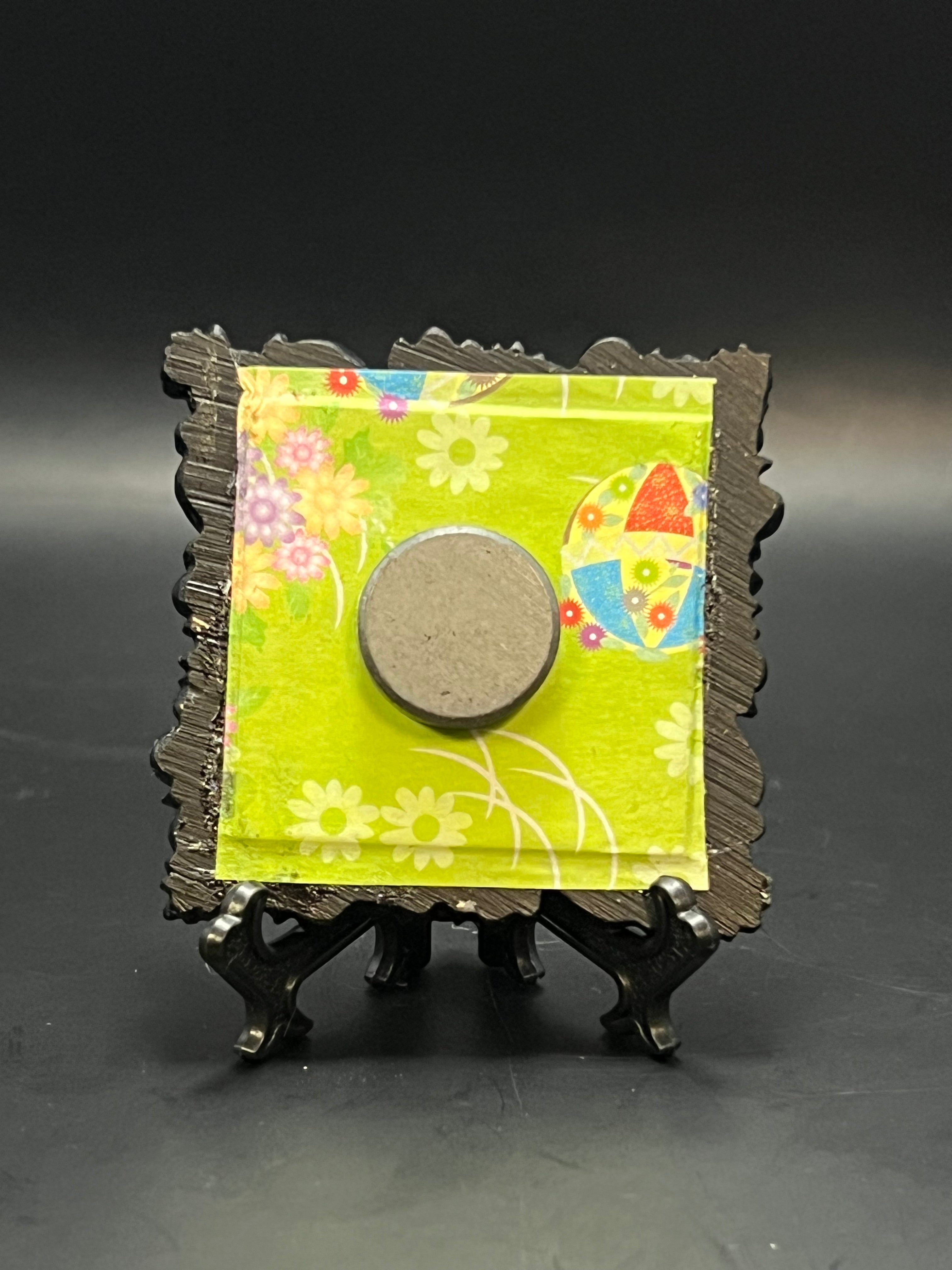 This is the handmade Fly Agaric Mushroom Miniature Painting by Chris Foster Design. This is showcasing the back. This is a small square frame and the decorative item with a green background with flowers and balls with red, white, yellow, orange and blue colors. There is a magnet and against a miniature picture frame.