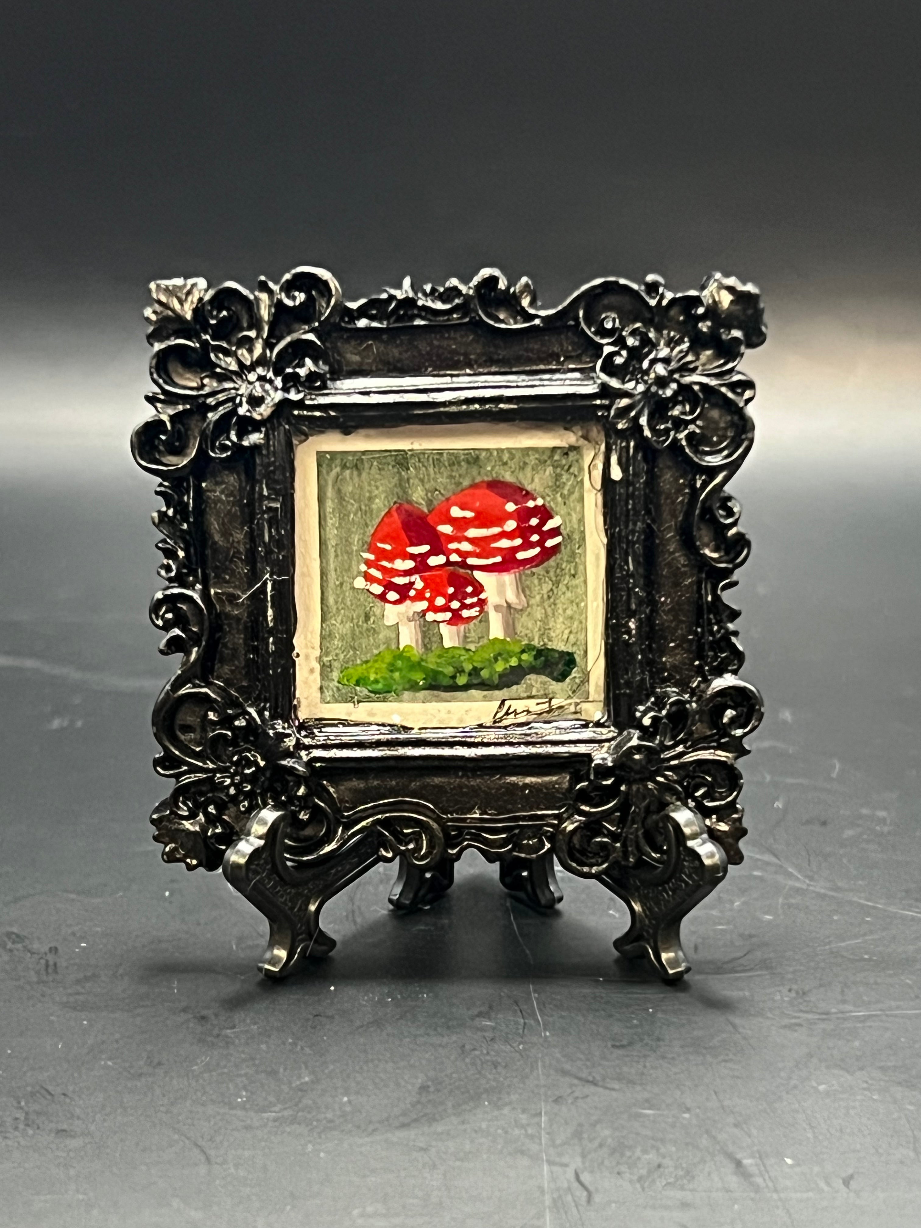 This is the handmade Fly Agaric Mushroom Green Miniature Painting by Chris Foster Design. This is showcasing the front. This is a decorative piece with a black square frame, there are three red and white speckled mushrooms in different sizes on top of dark green grass/moss. The background is a grey and green background. 