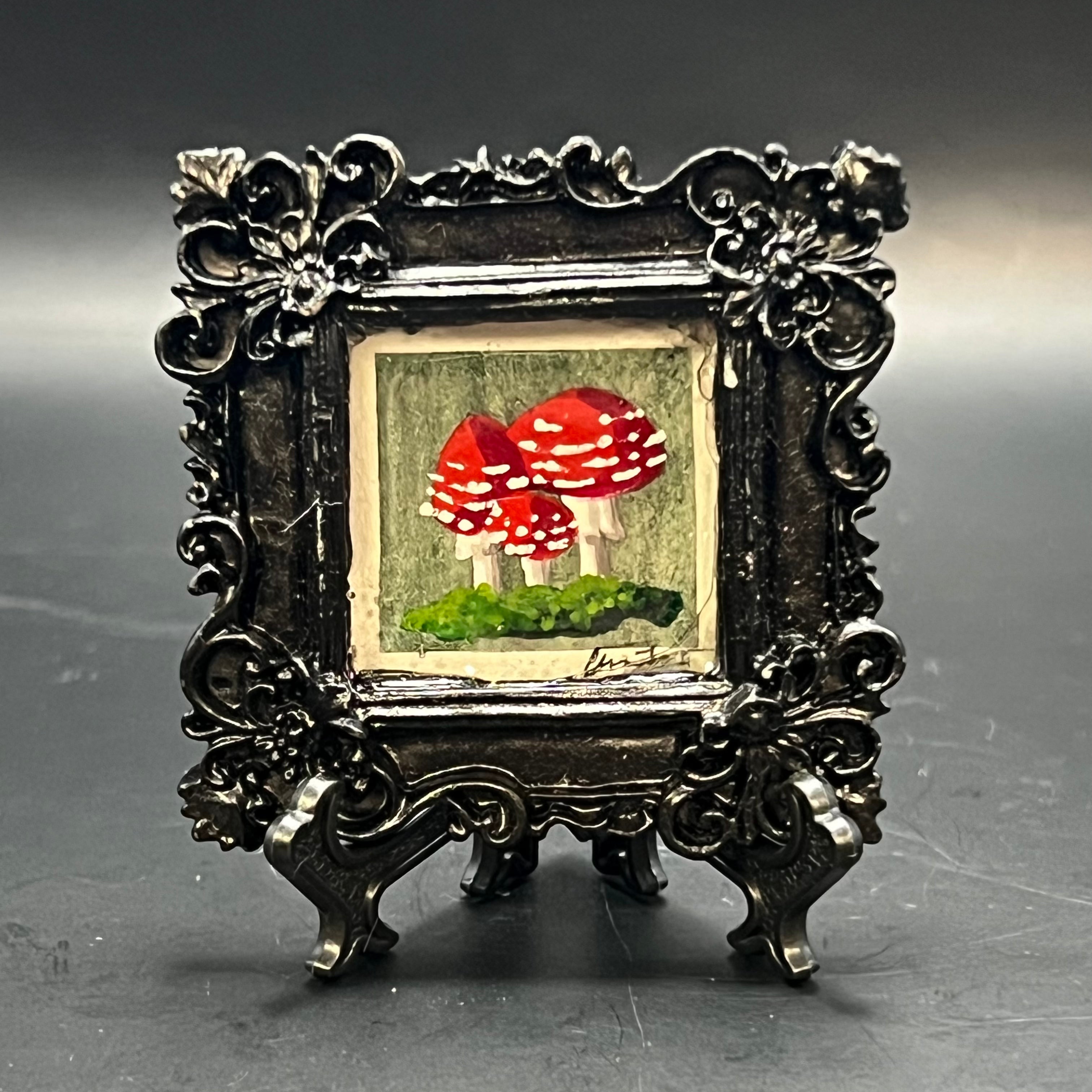 This is the Fly Agaric Mushroom Green Mini Painting by Chris Foster Design. This is showcasing the front. Ornamental gloss black square frame. Three red fly agaric mushrooms nestled in moss on a mossy green watercolor background. The painting is on a black stand and is against a dark background.