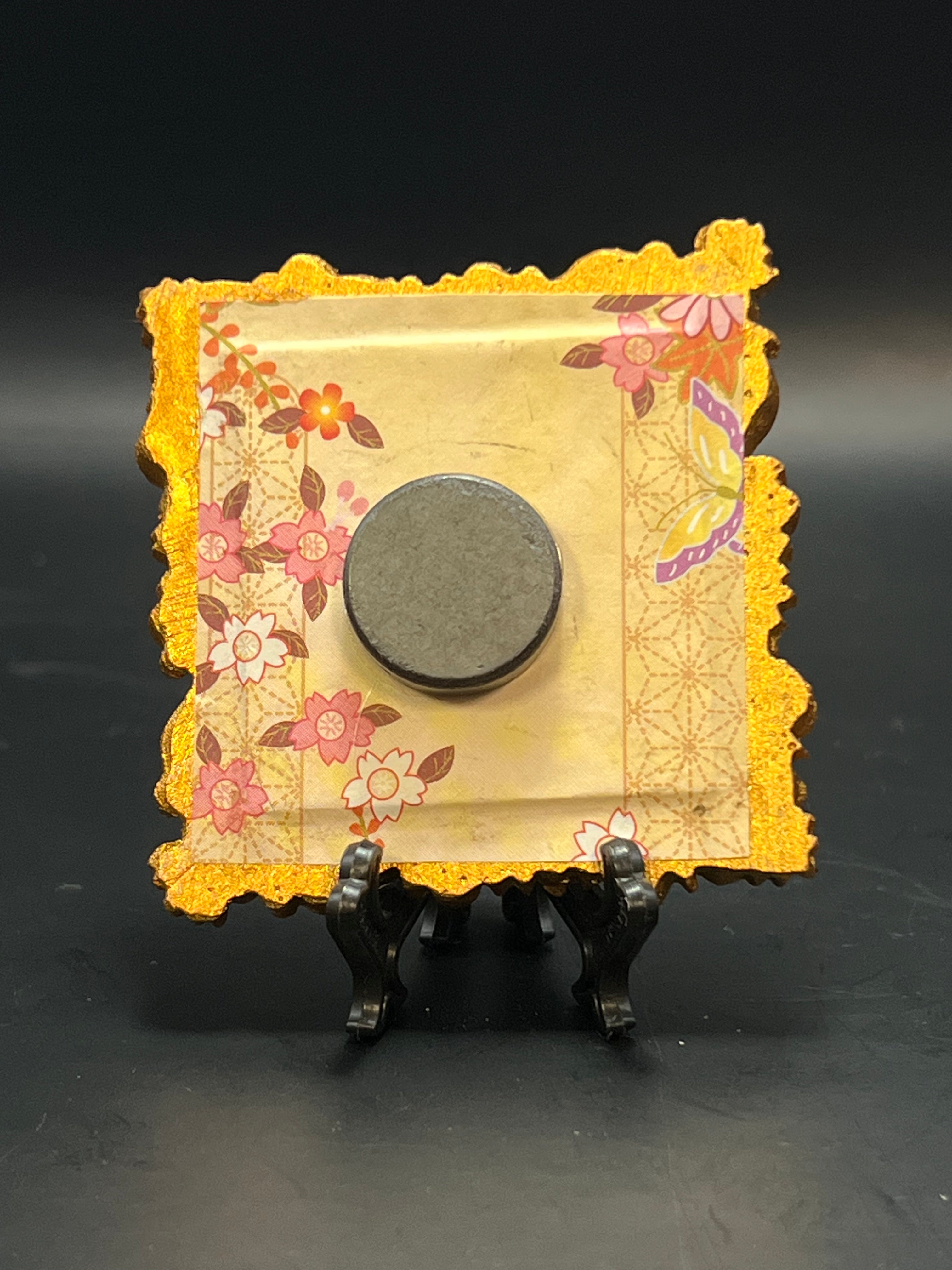 This is the handmade Fly Agaric Orange Miniature Painting by Chris Foster Design. This is showcasing the back. This is a small square frame and the decorative item with a yellow background with flowers falling on the right corner and left side. There is an intricate design on both sides. The flowers are red, white and yellow with maroon leaves. The pattern is maroon. There is a magnet and against a miniature picture frame.