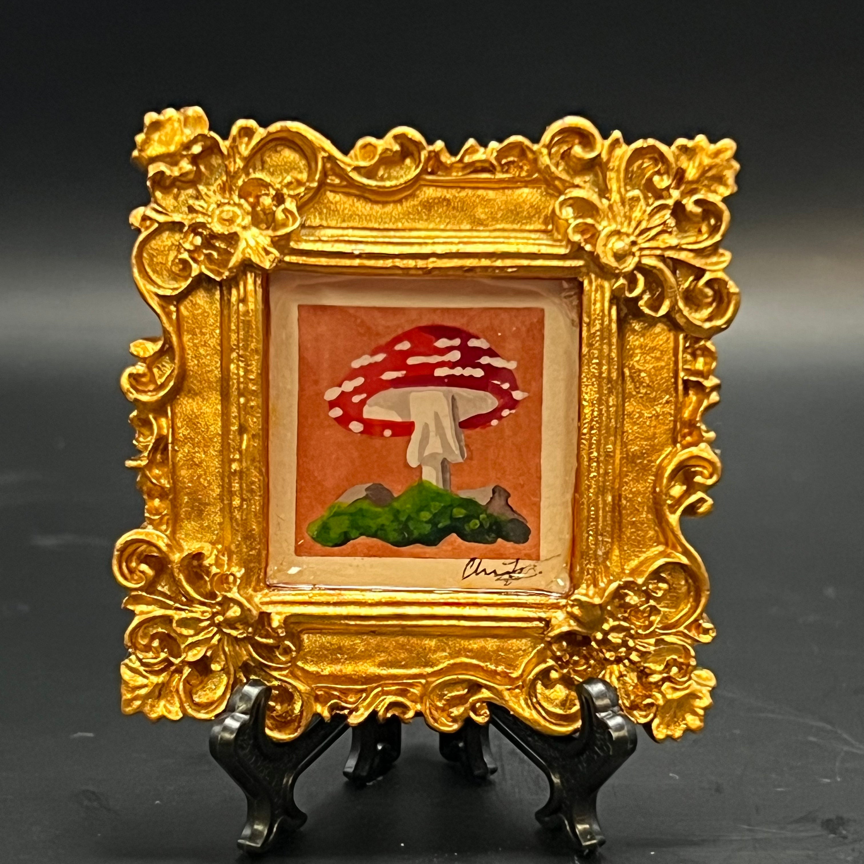 This is the Fly Agaric Orange Mini Painting by Chris Foster Design. This is showcasing the front. Ornate metallic gold square frame. One large red fly agaric sprouting from rocky moss on an orange watercolor background. It is against a dark background.