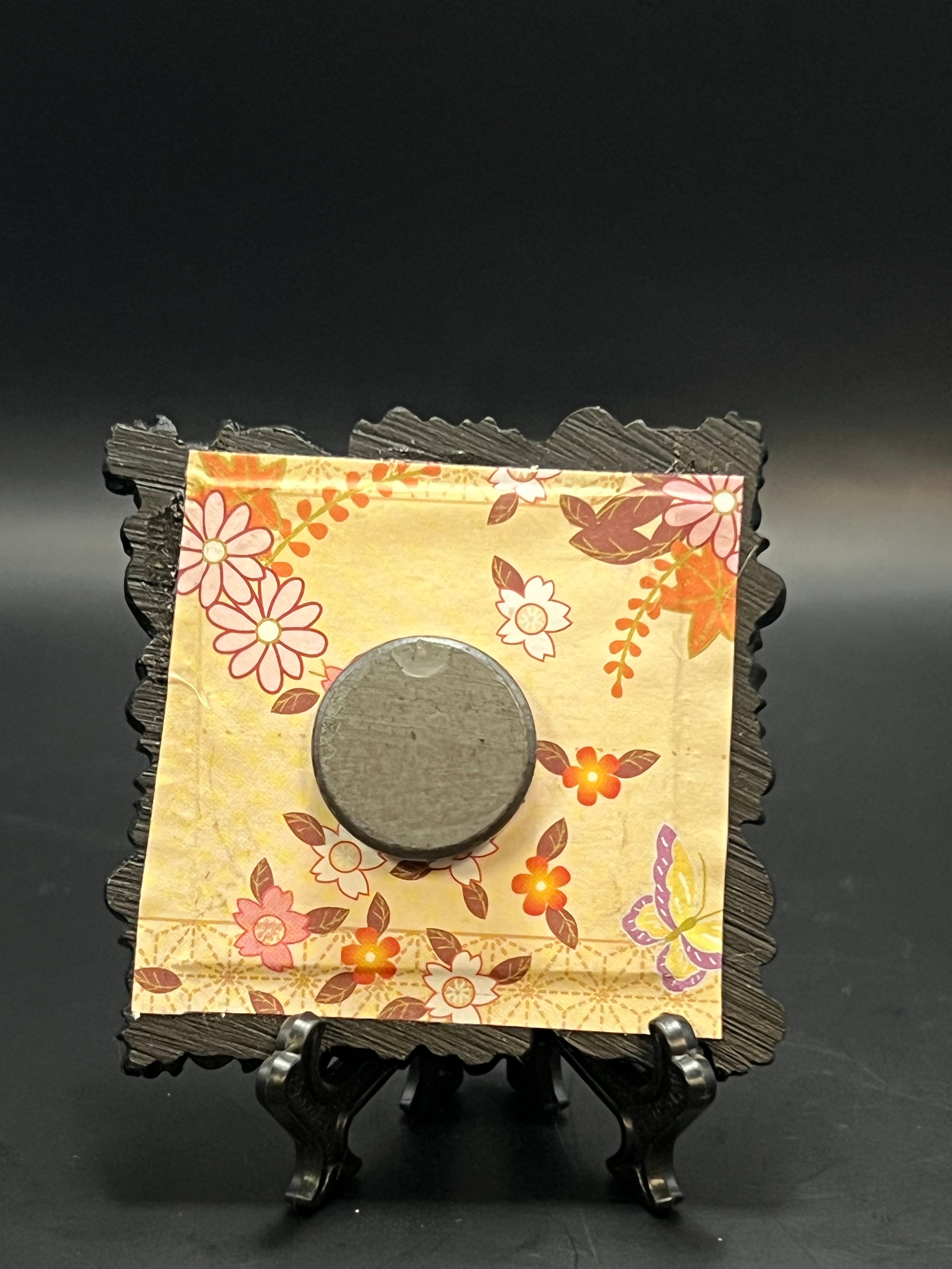 This is the handmade Fly Agaric Yellow Miniature Painting by Chris Foster Design. This is showcasing the back. This is a small square frame and the decorative item with a yellow background containing flowers and leaves throughout. The flowers are white, red and yellow with orange and maroon leaves. There is a magnet and against a miniature picture frame.