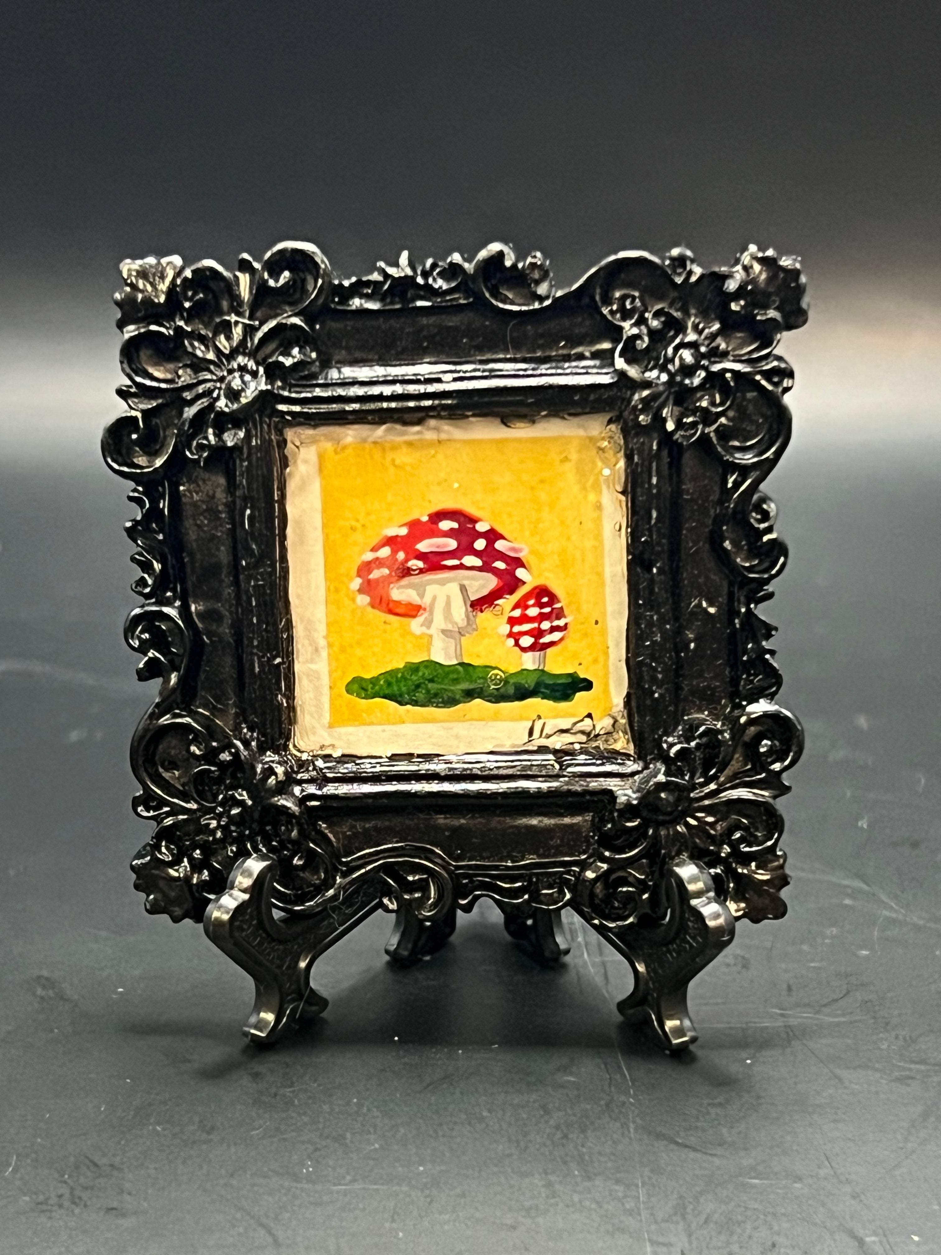 This is the Fly Agaric Yellow Miniature Painting by Chris Foster Design. This is a decorative squared black frame with a small painting of two red and speckled white mushrooms on a yellow background resting on dark green moss/grass. The frame is on a stand.