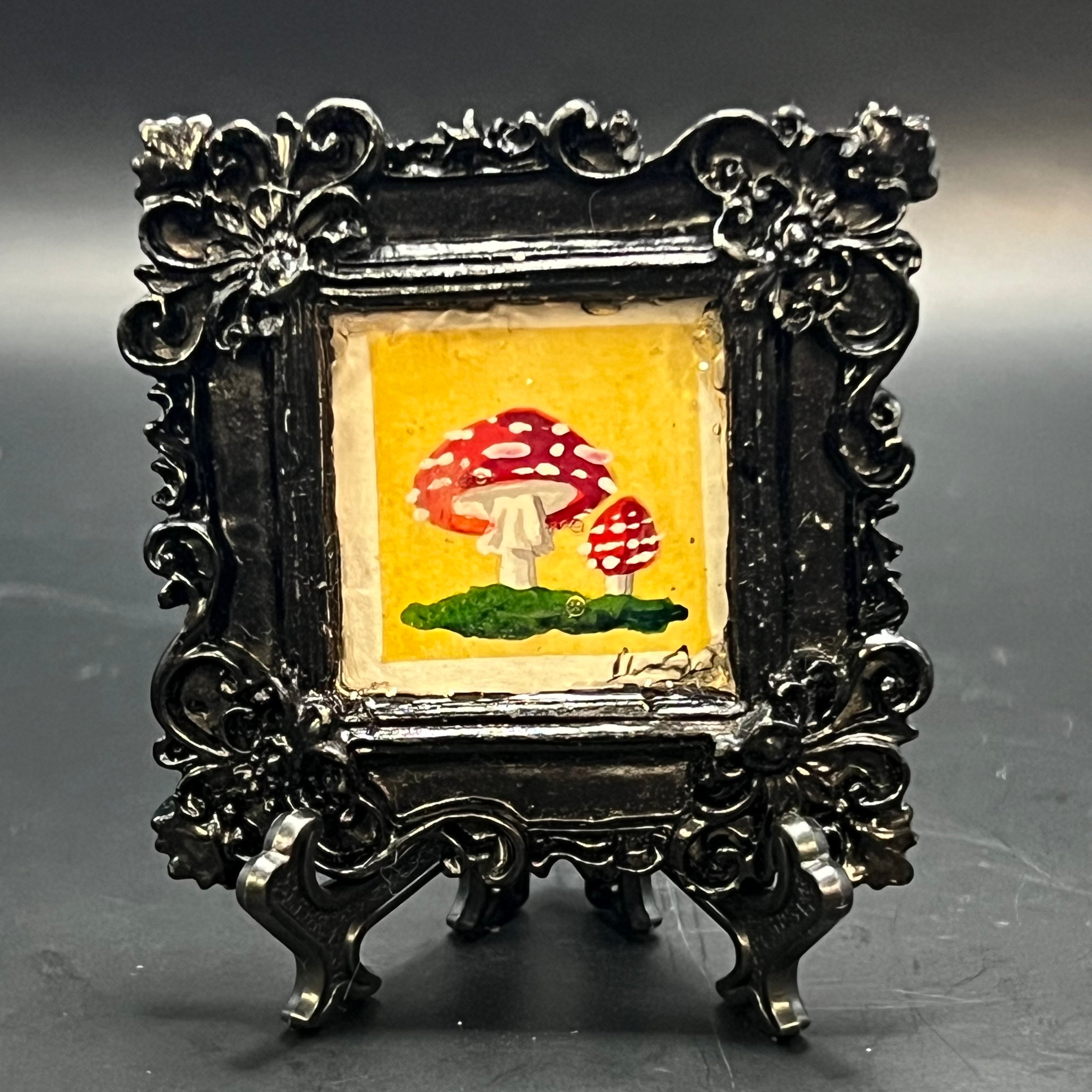 This is the Fly Agaric Yellow Mini Painting by Chris Foster Design. This is showcasing the front. Ornate Glossy Black square frame. Two red Fly agaric mushrooms imbedded in green moss on a yellow watercolor background. It is against dark background.