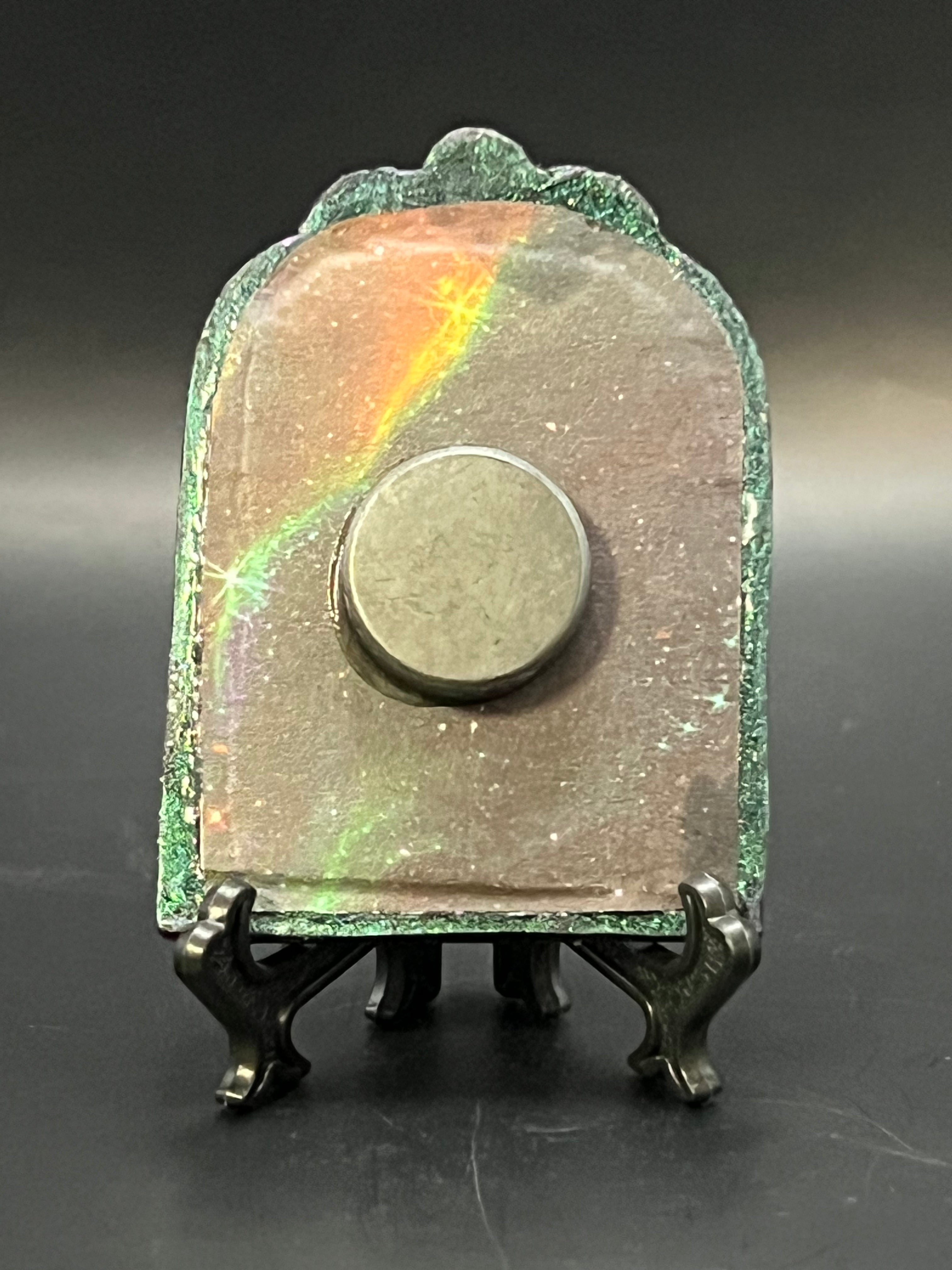 This is the handmade Gem in the Void Miniature Painting by Chris Foster Design. This is showcasing the back. Decorative piece with a metallic base on a dark background on a stand. There is a magnet and against a miniature picture frame.