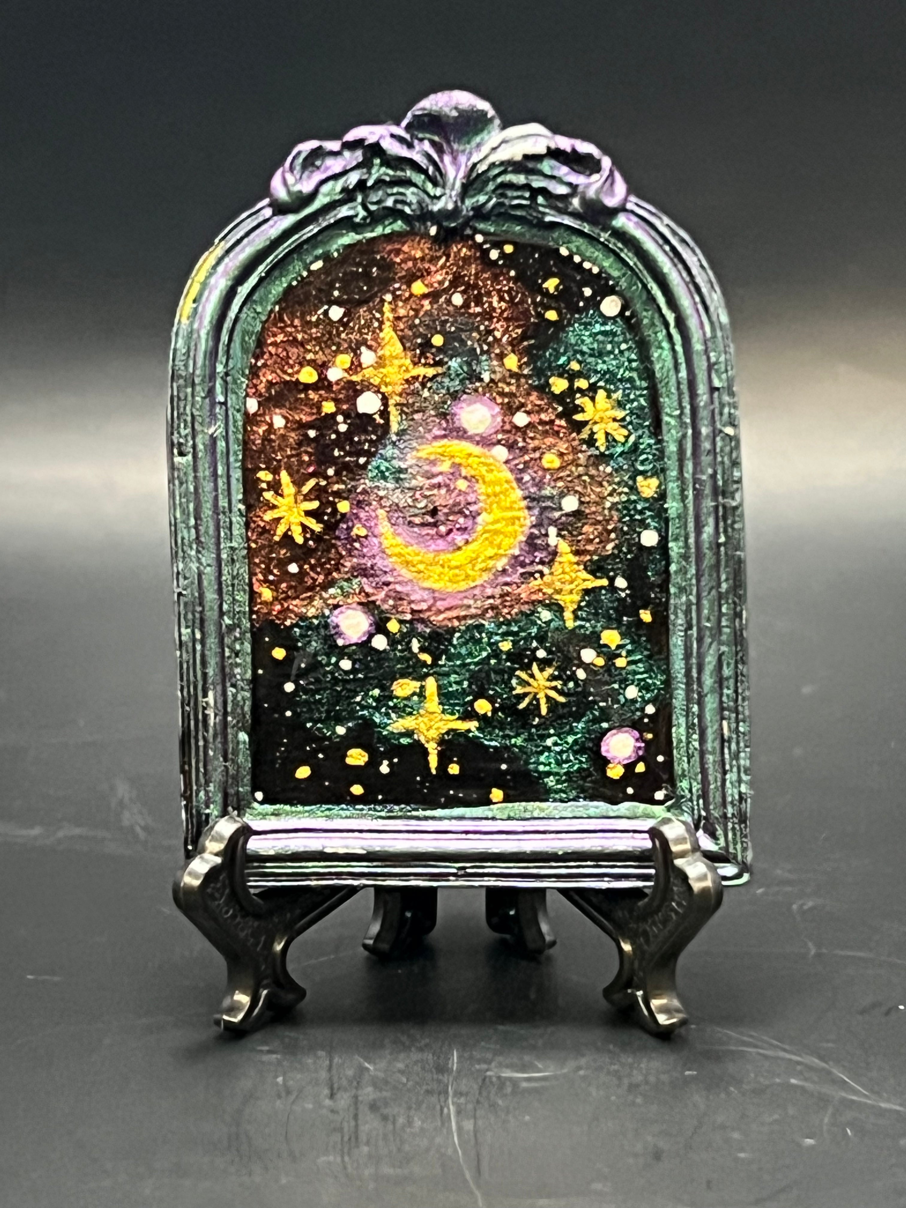 This is the handmade Gem in the Void Miniature Painting by Chris Foster Design.This is showcasing the front. It is a decorative piece with a celestial design featuring stars and a moon on a stand against a dark background. The colors are sparkling yellow, orange, green and black that swirl around. There is a magnet and against a miniature picture frame.
