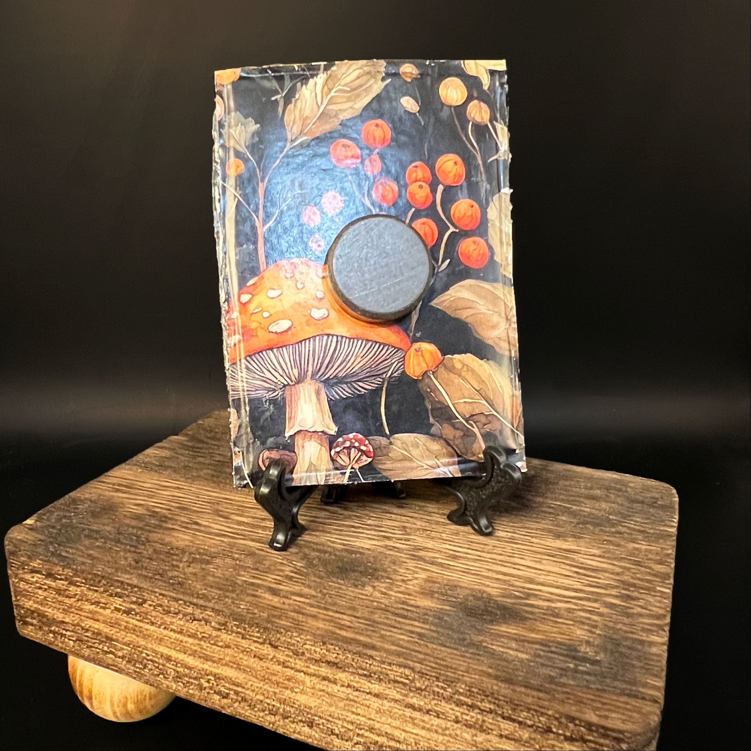 This is the Ghost Mini Painting by Chris Foster Design. This is showcasing the back of the painting. The painting has a black rectangular frame with a round magnet in the middle. The painting is backed with a mushrooms and berries design on a wooden stand against a dark background.