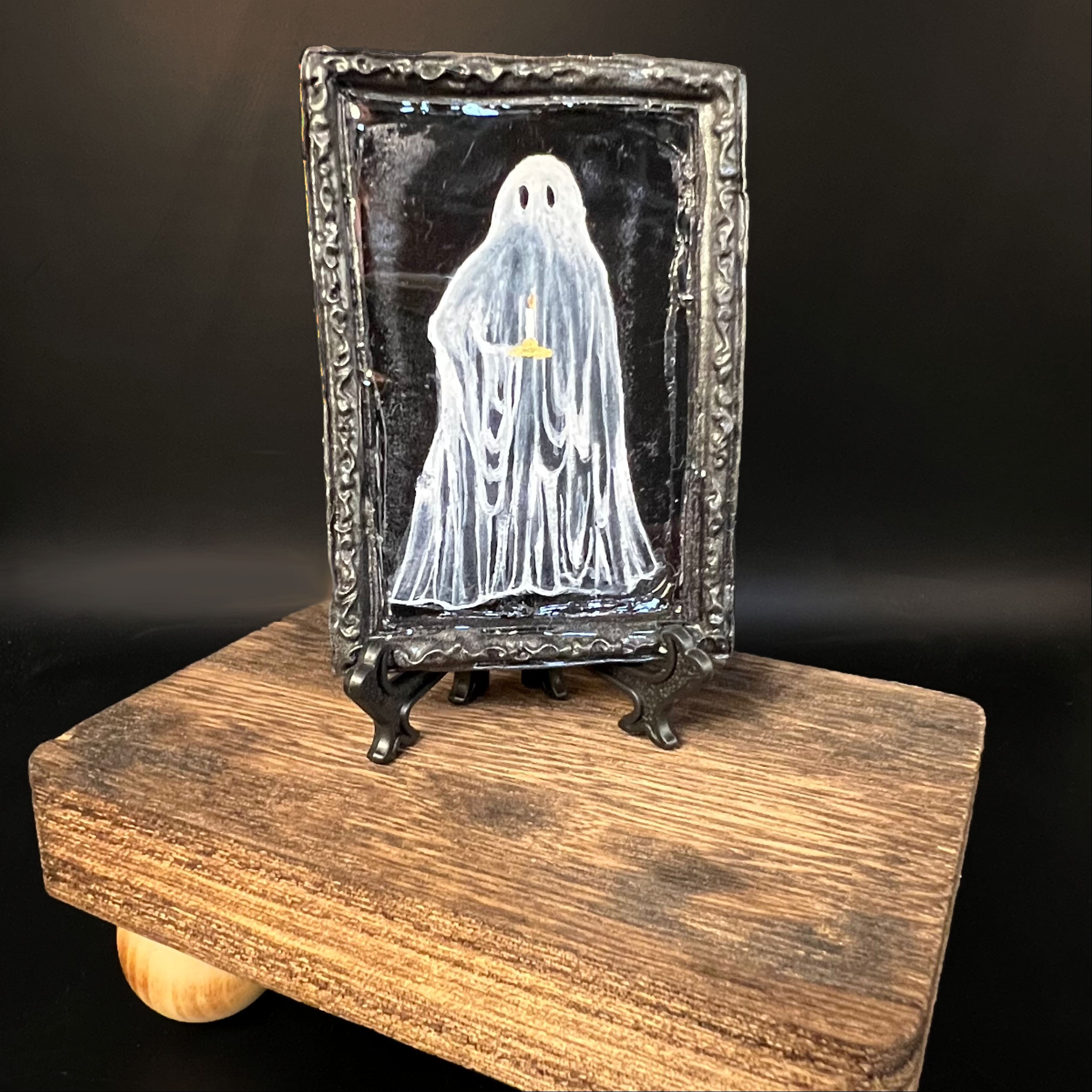 This is the Ghost Mini Painting by Chris Foster Design. This is showcasing the front of the painting. The painting has a black rectangular frame with a white transcluscent ghost in the middle holding a lit candle. The background of the painting is black and on a black stand. The miniature and stand is on a wooden board against a dark background. 