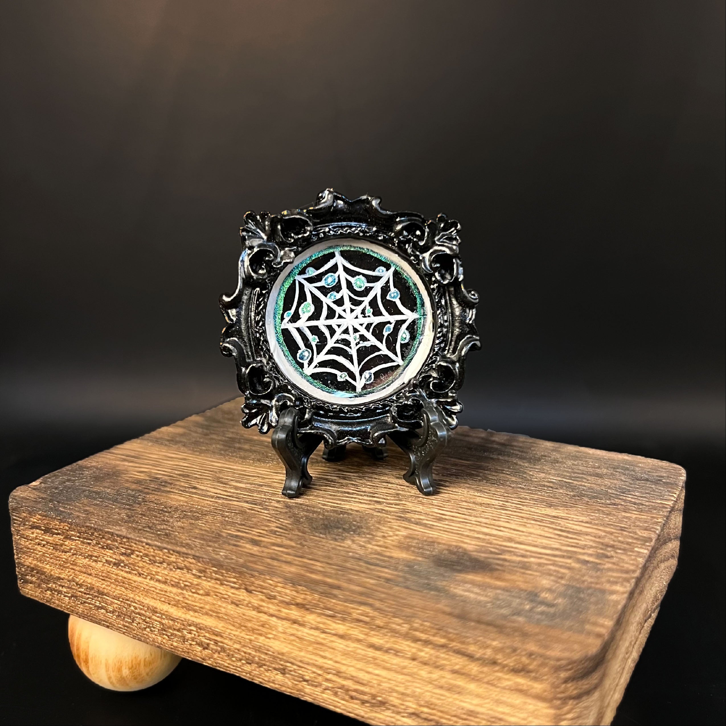 This is the Glistening Web Mini Painting by Chris Foster Design. This is showcasing the front of the painting. The painting has a black circular frame with a spider web in white, shimmery blue water droplets design against a black background. There is a shimmery blue outline for the illustration, on a black stand. The miniature painting in general is on a wooden stand against a dark background.