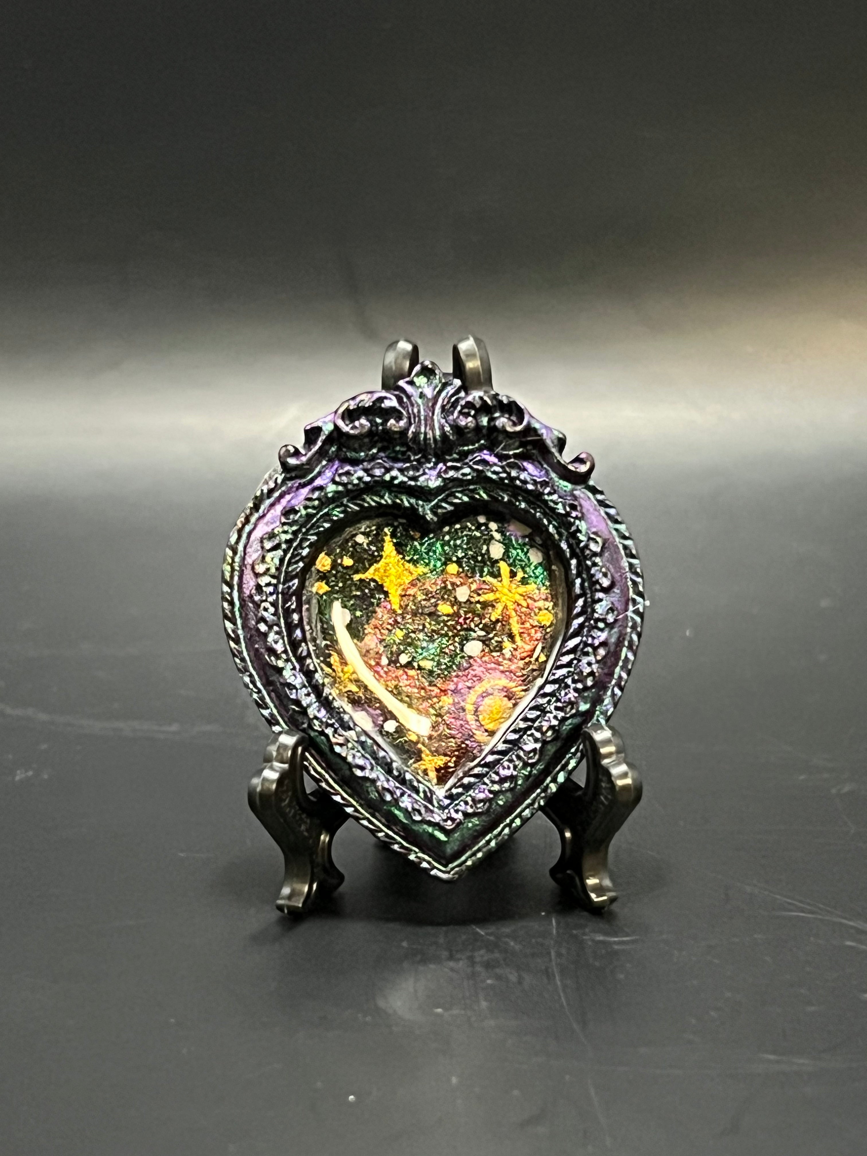 This is the handmade Glowing Cluster Miniature Painting by Chris Foster Design. This is showcasing the front. This is a decorative piece with a shimmery black/purple heart frame, starry night design including large yellow stars, small white stars, sparkling green, orange, yellow and black on a dark background. There is a magnet and against a miniature picture frame.