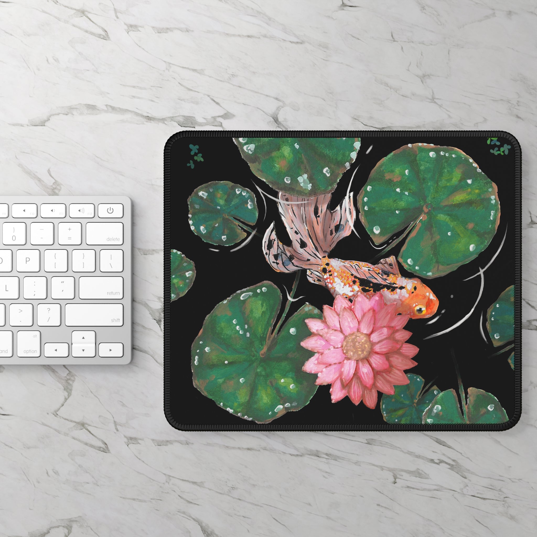This is the Goldfish Mouse Pad by Chris Foster Design. This shows the front corner where the threading begins. The illustration has the background in black, nine dark or light green lily pads, small foliage, one orange, white and black goldfish and a pink blooming flower. The mouse pad is on a marbled silver/white surface with a white and silver keyboard to the left. 