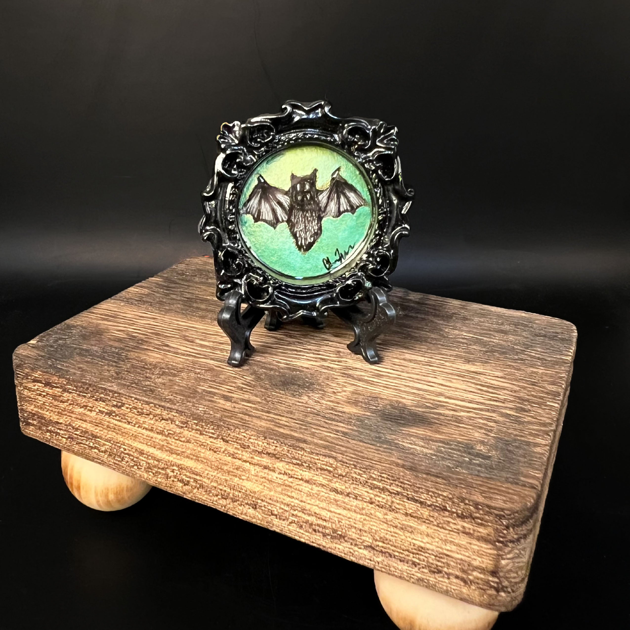 This is the Green Bat Mini Painting by Chris Foster Design. This is showcasing the front of the painting. The painting has a black circular frame with a black bat design on a gradient green background. The studio's signature on the right, on a black stand and all together on a wooden stand against a dark background.