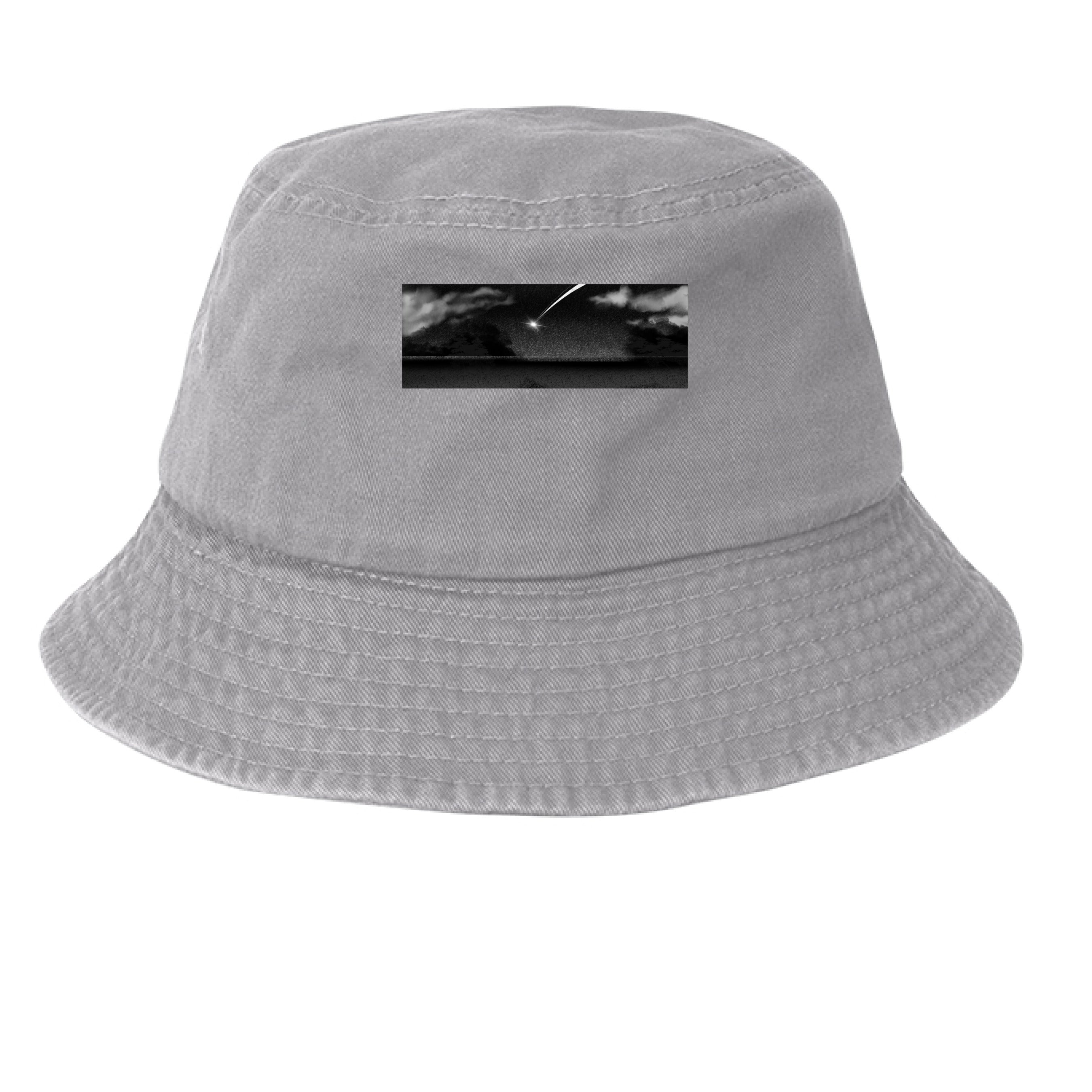 This is the Grey Star Fall Bucket Hat by Lee Hansheng Studios. This is showcasing the hat from the front. The illustration is a meteor streaking through the sky. It is a moody piece that as gradients of black and grey, a little pixelated with white clouds in the forefront. The meteor is also white. It is a smaller rectangular than the cap. It is against a white background.