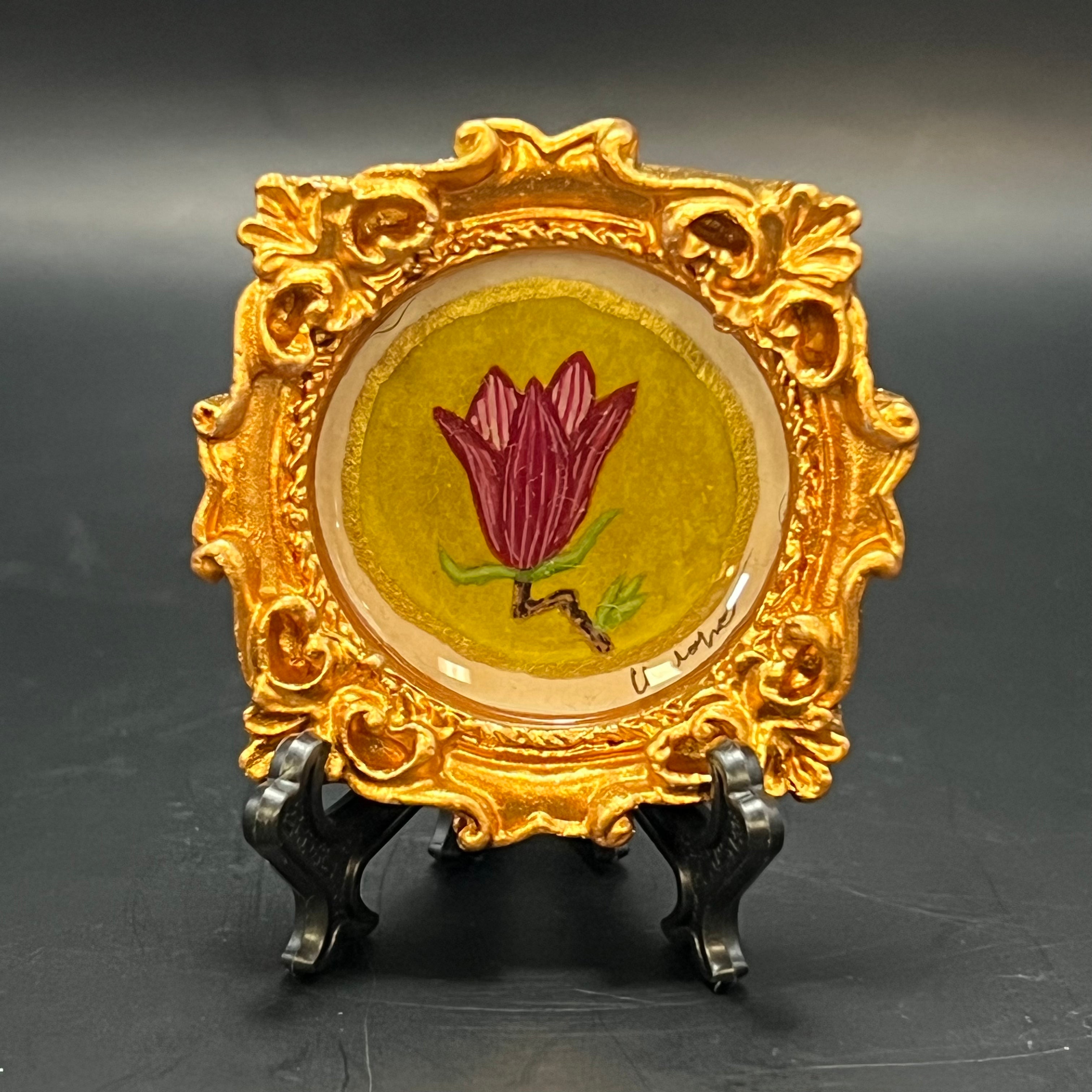This is the Japanese Magnolia Blossom Mini Painting by Chris Foster Design. This is showcasing the front. Ornate golden circle frame. This is the magenta Japanese magnolia bloom with delicate branch and green leaves on bright green gold washed background. It is against a dark background.