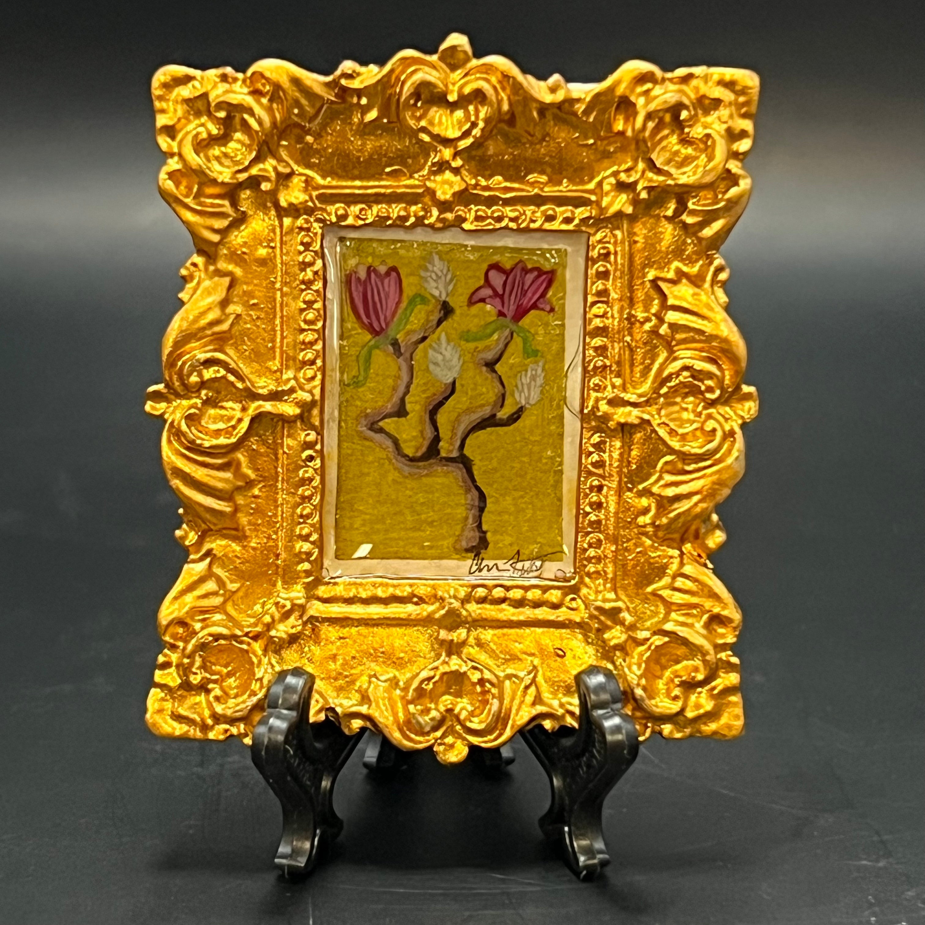 This is the Japanese Magnolia Double Bloom Mini Painting by Chris Foster Design. This is showcasing the front. There's an ornate olden rectangular frame. Double blooming magenta japanese magnolia branch on a bright green background with a gold glitter wash.  It is against a dark background.
