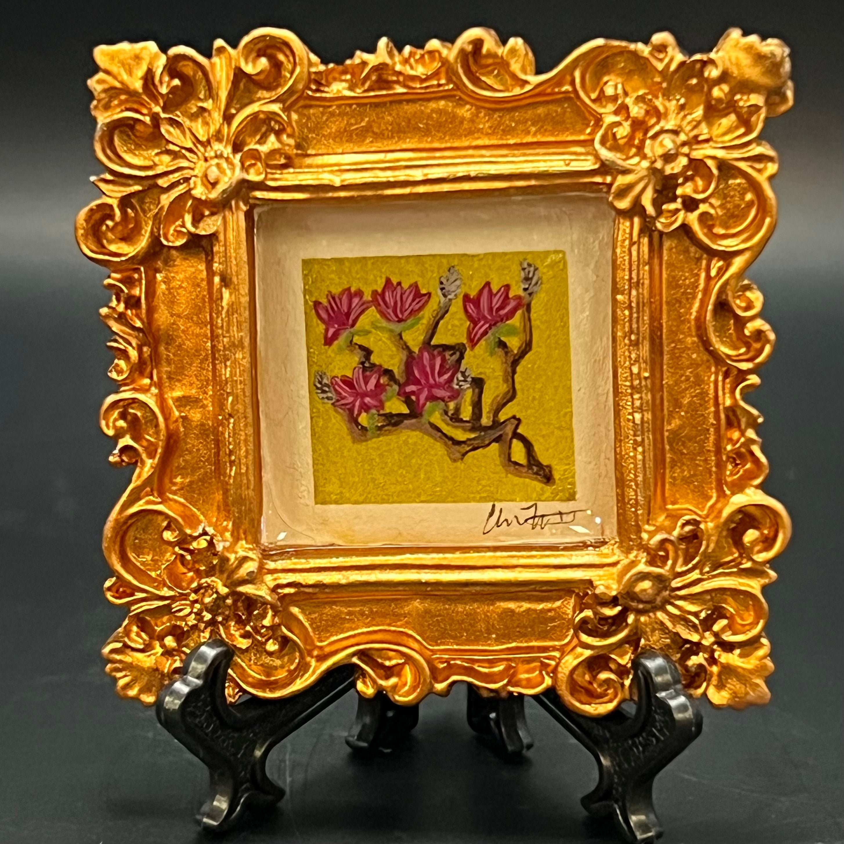 This is the Japanese Magnolia Mini Painting by Chris Foster Design. The ornate gold frame is encasing the painting. The illustration is of branching magenta japanese magnolia painting on bright green background with gold glitter wash. The painting is on a stand against a dark background.
