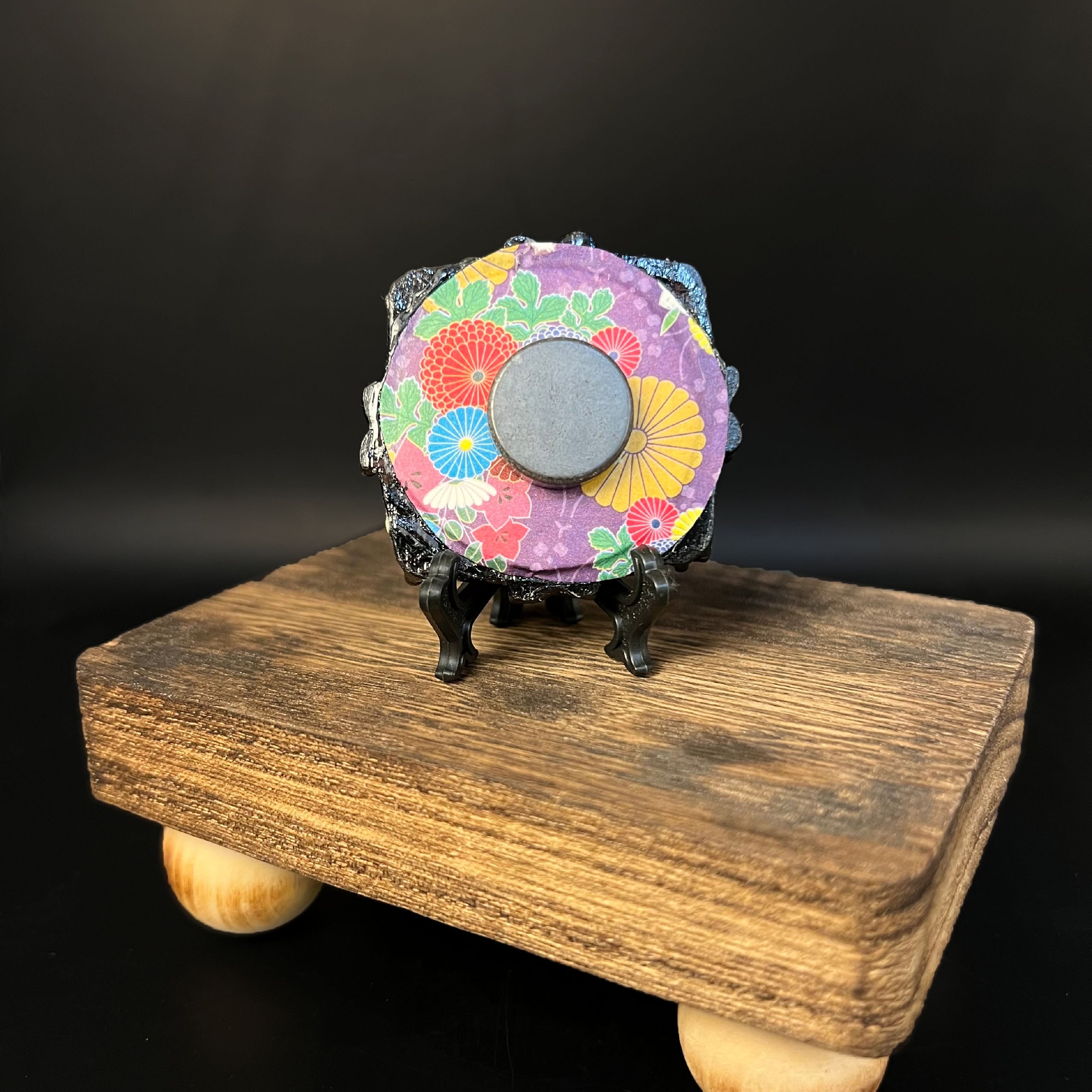 This is the Lantern Mini Painting by Chris Foster Design. This is showcasing the back of the painting. The painting has a circular black decorative frame with a round magnet. The backing is a floral design with yellow, red, blue, green leaves, light purple flowers and purple background. The miniature painting is on a wooden stand against a black background.