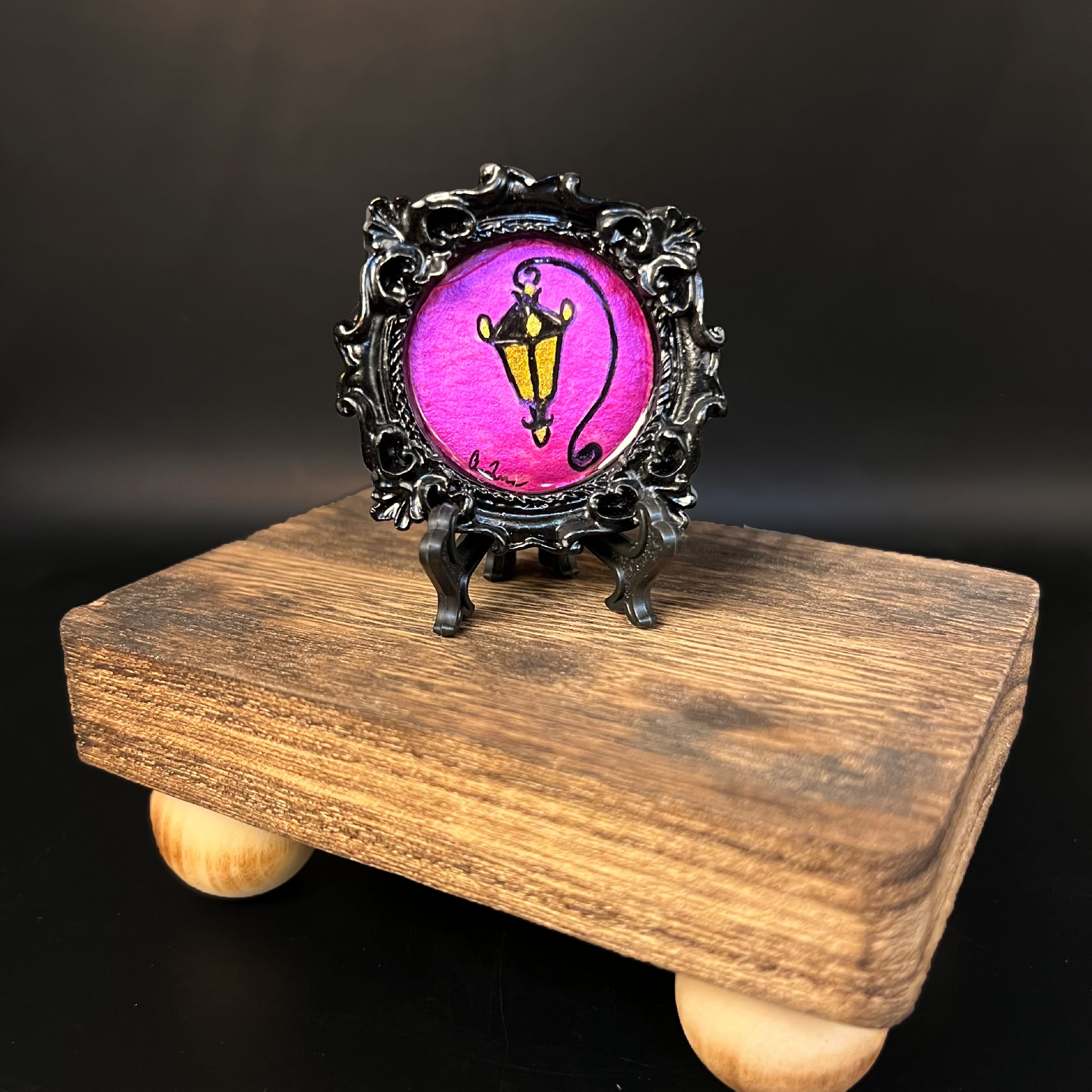 This is the Lantern Mini Painting by Chris Foster Design. This is showcasing the front of the painting. The painting has a circular black decorative frame with a pink circular design. The design is of an old time lamp that is lit with yellow, the studio's signature on the left and on a wooden stand against a black background.
