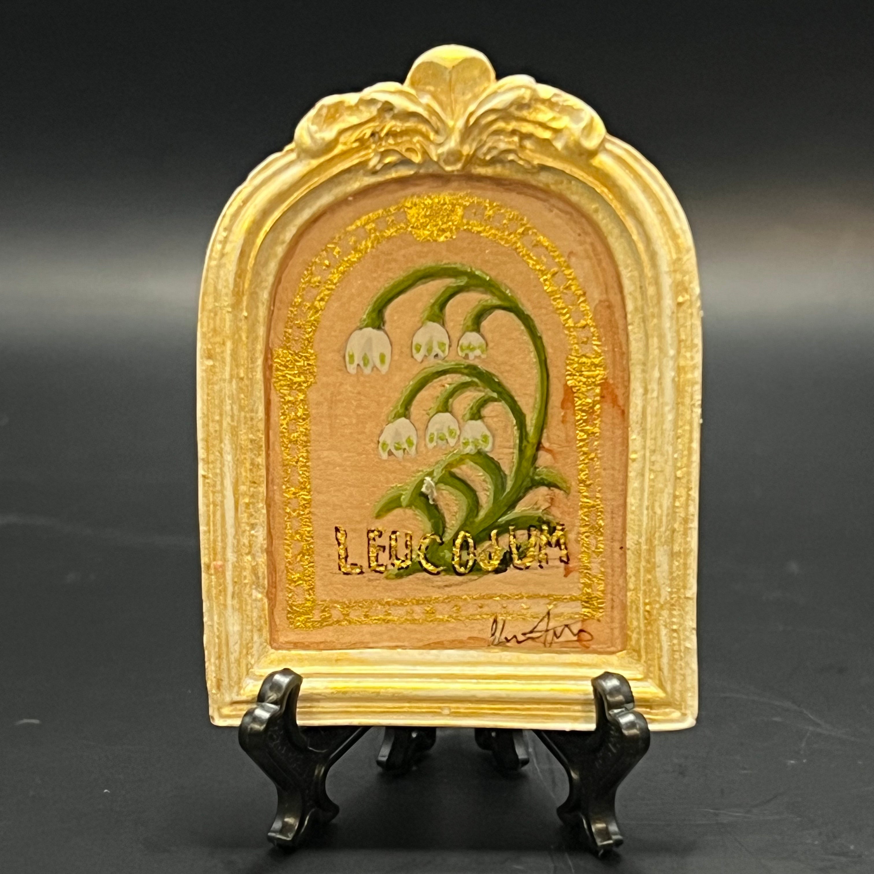 This is the Leucojum Mini Painting by Chris Foster Design. This is showcasing the front. Ornate white round top frame with gold distress. Leucojum painting with white flowers green stems and leaves on a nude gold glitter background with gold border. It is against a dark background.