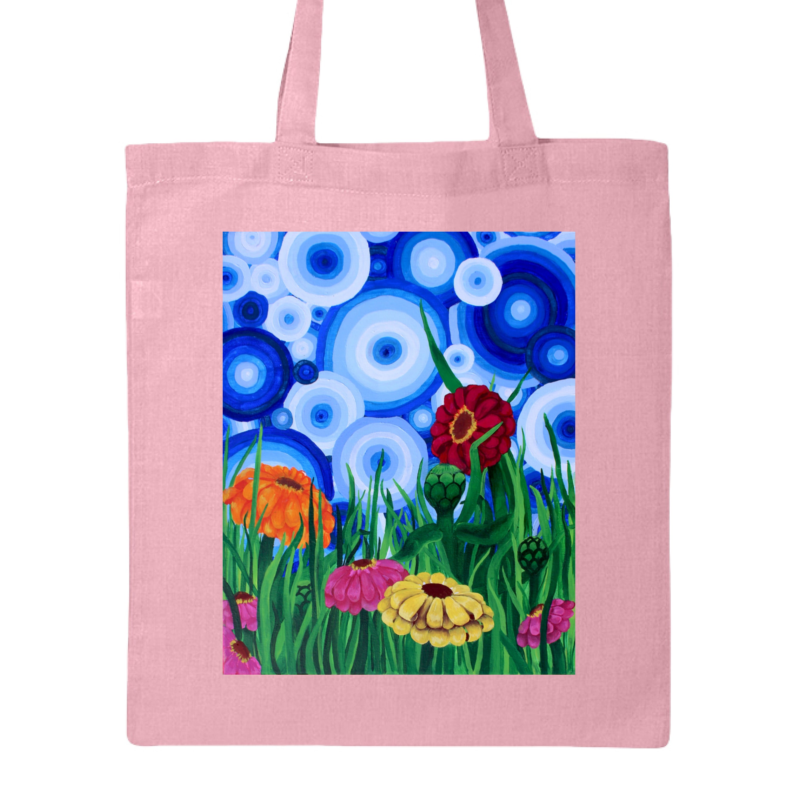 This is the Light Pink Zinnia 2 Canvas Tote by Storm Garden Studio. This showcases the front of the tote. The rectangular illustration are bold and color zinnias like red, yellow and orange across tall green grass. The background has many different circles in blue, dark blue and white. The tote is against a white background.