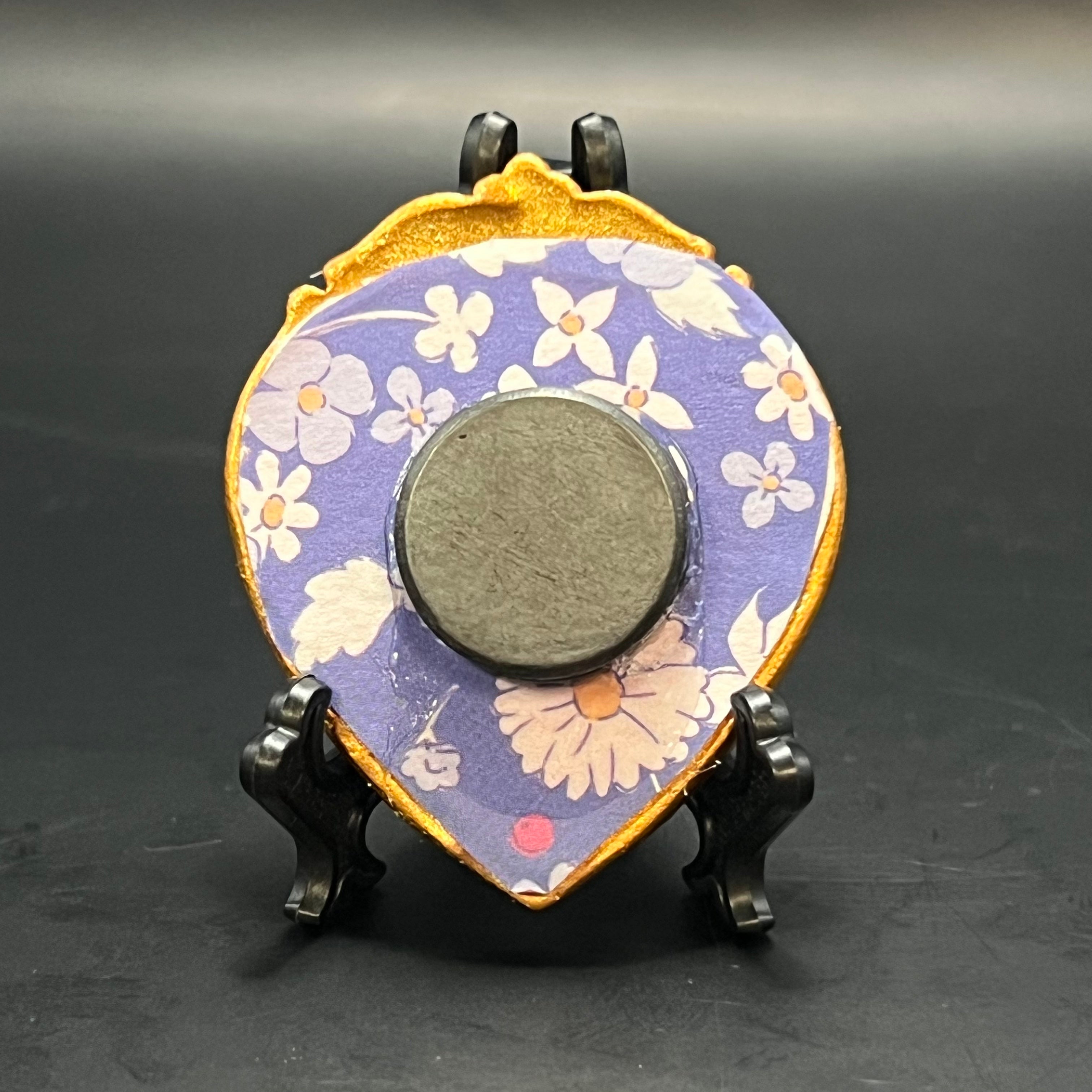 This is the Lilly of the Valley Mini Painting by Chris Foster Design. Ornate golden heart frame with purple and white floral design on a stand against a dark background.