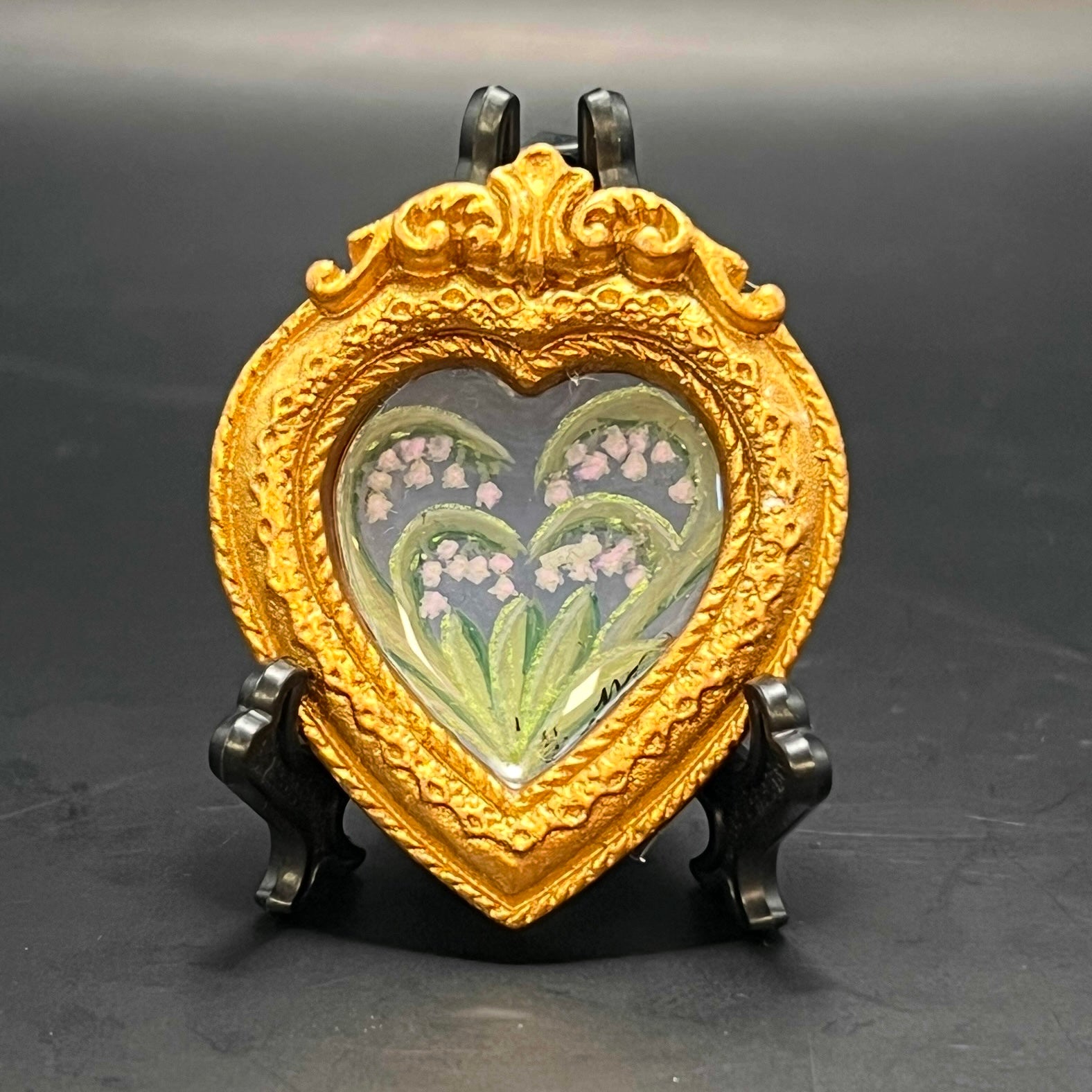 This is the Lilly of the Valley Mini Painting by Chris Foster Design. Ornate golden heart frame. Lily of the valley painting on light blue background with opalescent flowers, and metallic green accents on stems and leaves. Backed with blue floral pattern with yellow and red accents. It is against a dark background and is on a black stand. 