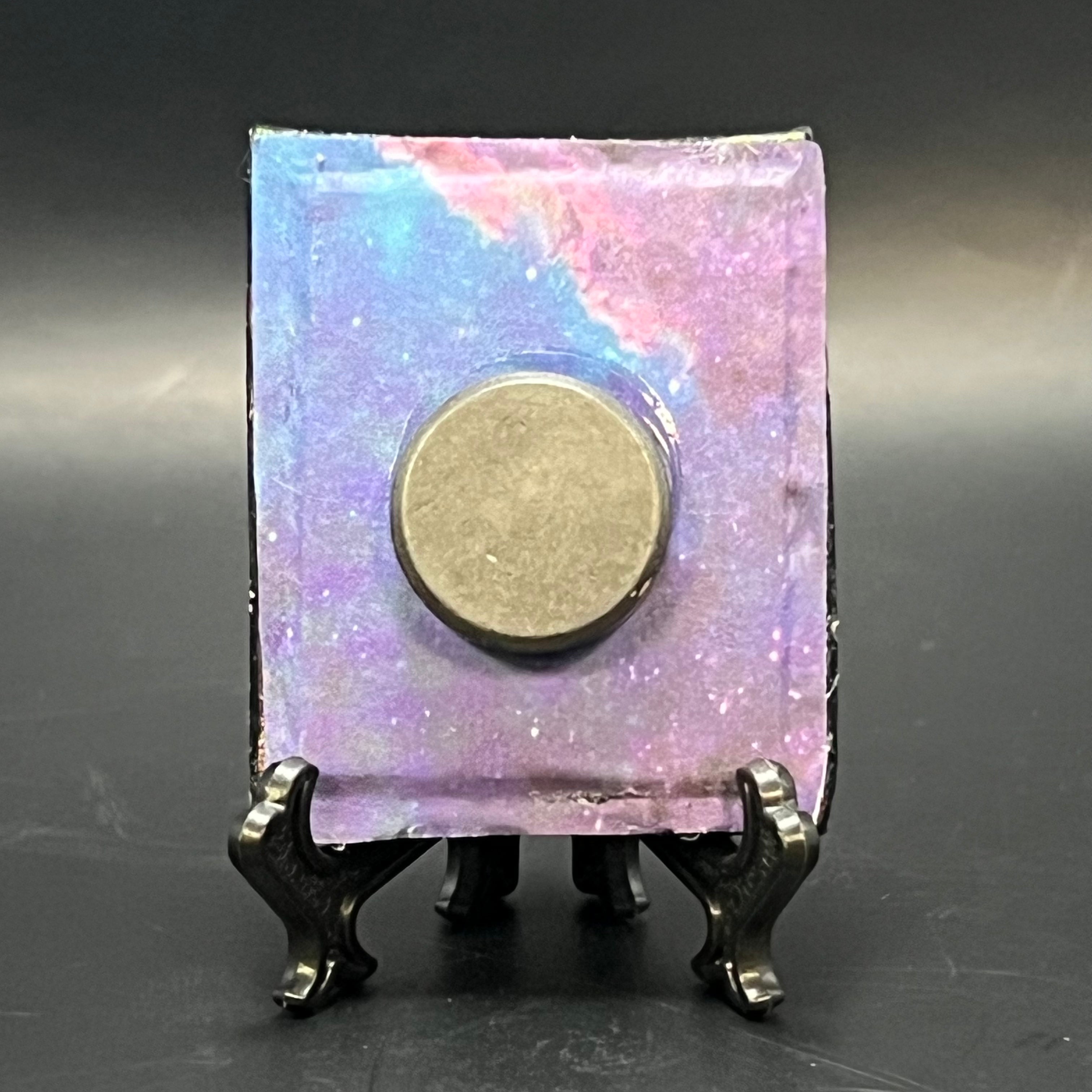 This is the Mii Spiral Galaxy Mini Painting by Chris Foster Design. This is showcasing the back. The backing is galaxy themed with colors of pink, blue and purple. In the middle is a round magnet, on a black stand and against a dark background.