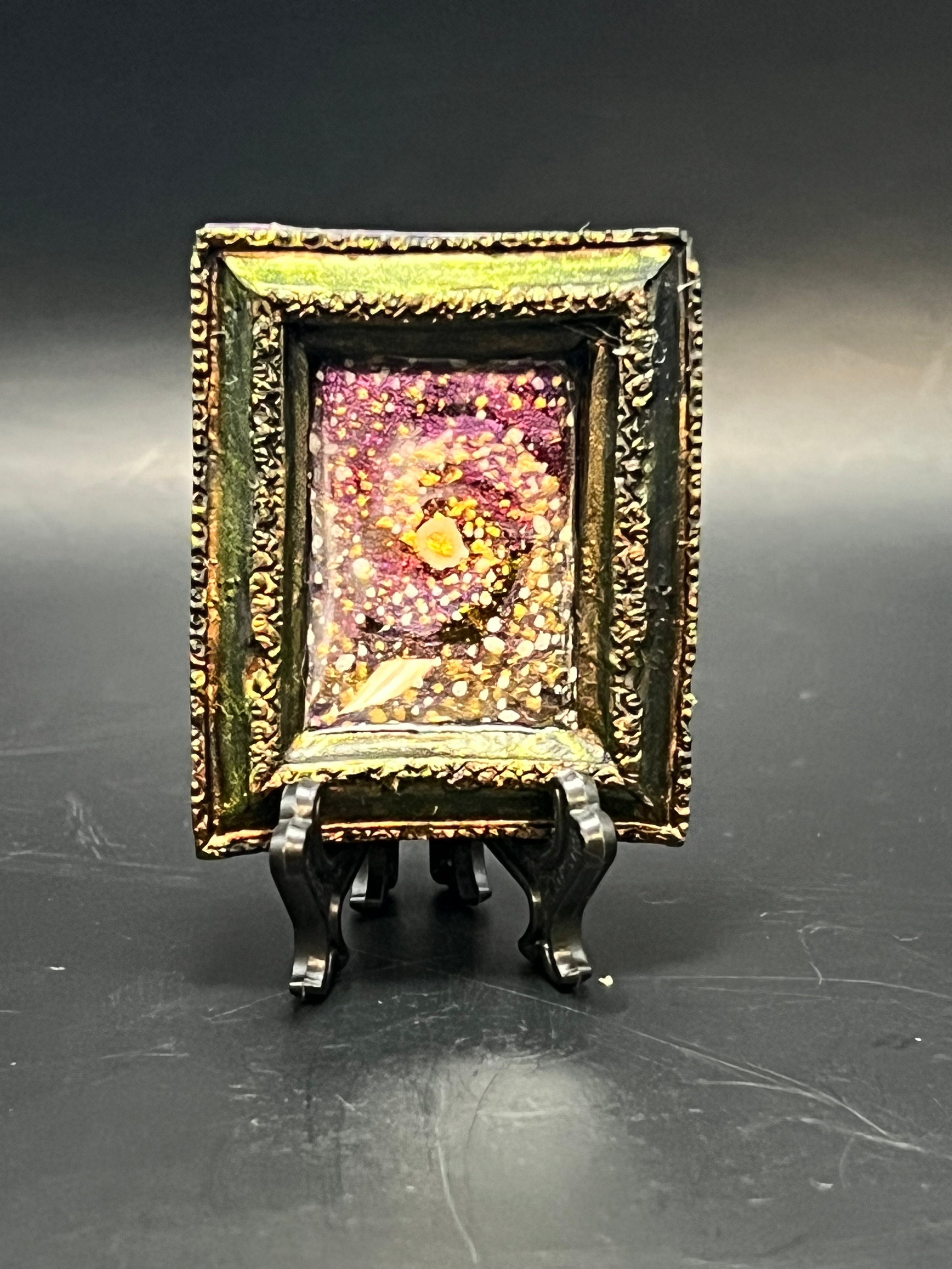 This is the handmade Mii Spiral Galaxy Miniature Painting by Chris Foster Design. This is showcasing the front. This is a decorative piece with a gold rectangular frame, spiral design including yellow stars, sparkling red, orange, yellow and white on a dark background. There is a magnet and against a miniature picture frame.