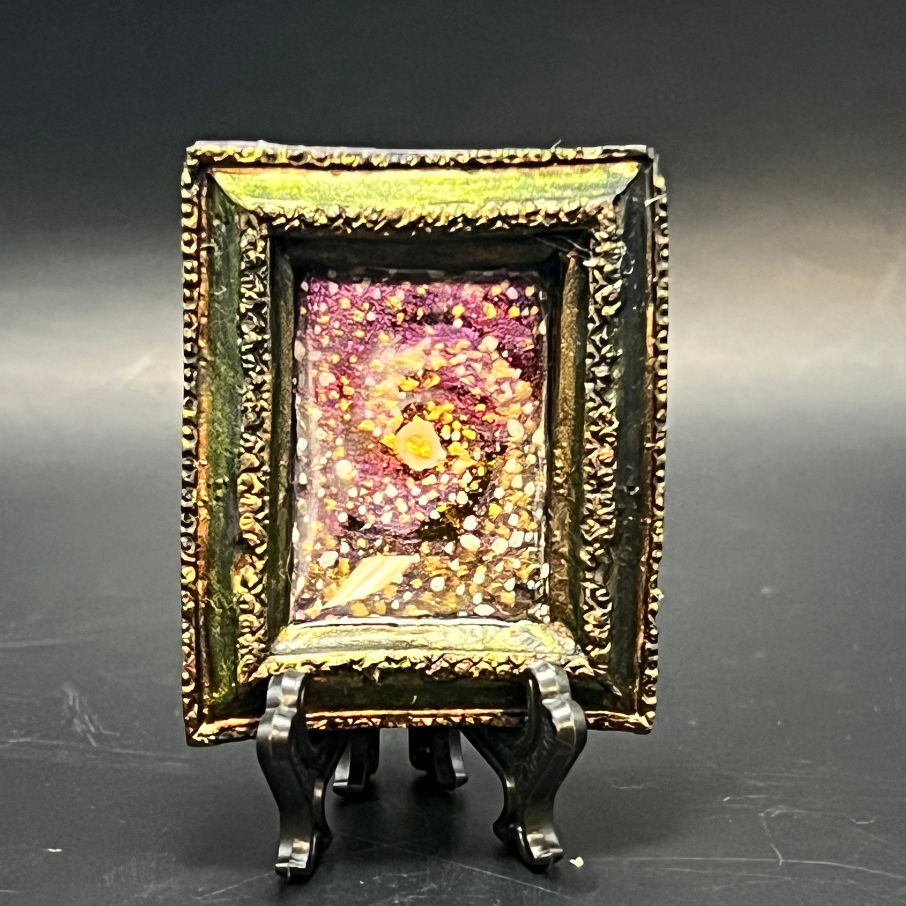 This is the Mii Spiral Galaxy Mini Painting by Chris Foster Design. This is showcasing the front. There is a decorative rectangular frame, gold and green sheer. The illustration is a spiral oof gold, background of pink, yellow and black that swirls around. The painting is on a black stand and is against a dark background.