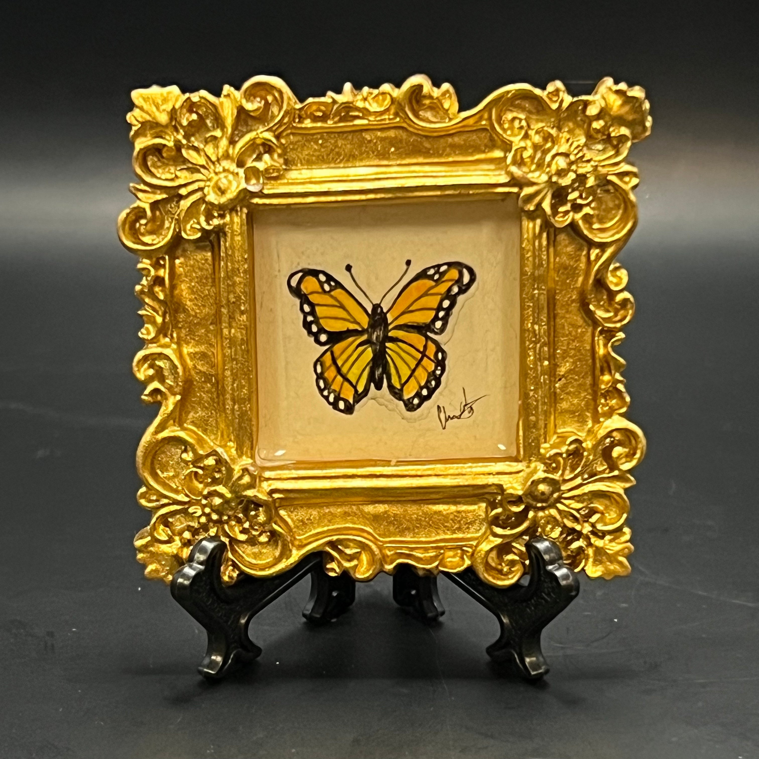 This is the Monarch Butterfly Mini Painting by Chris Foster Design. This is showcasing the front. Metallic gold ornate square frame. Orange monarch butterfly with black outline on a pale blue background. The painting is on a black stand and against a dark background.