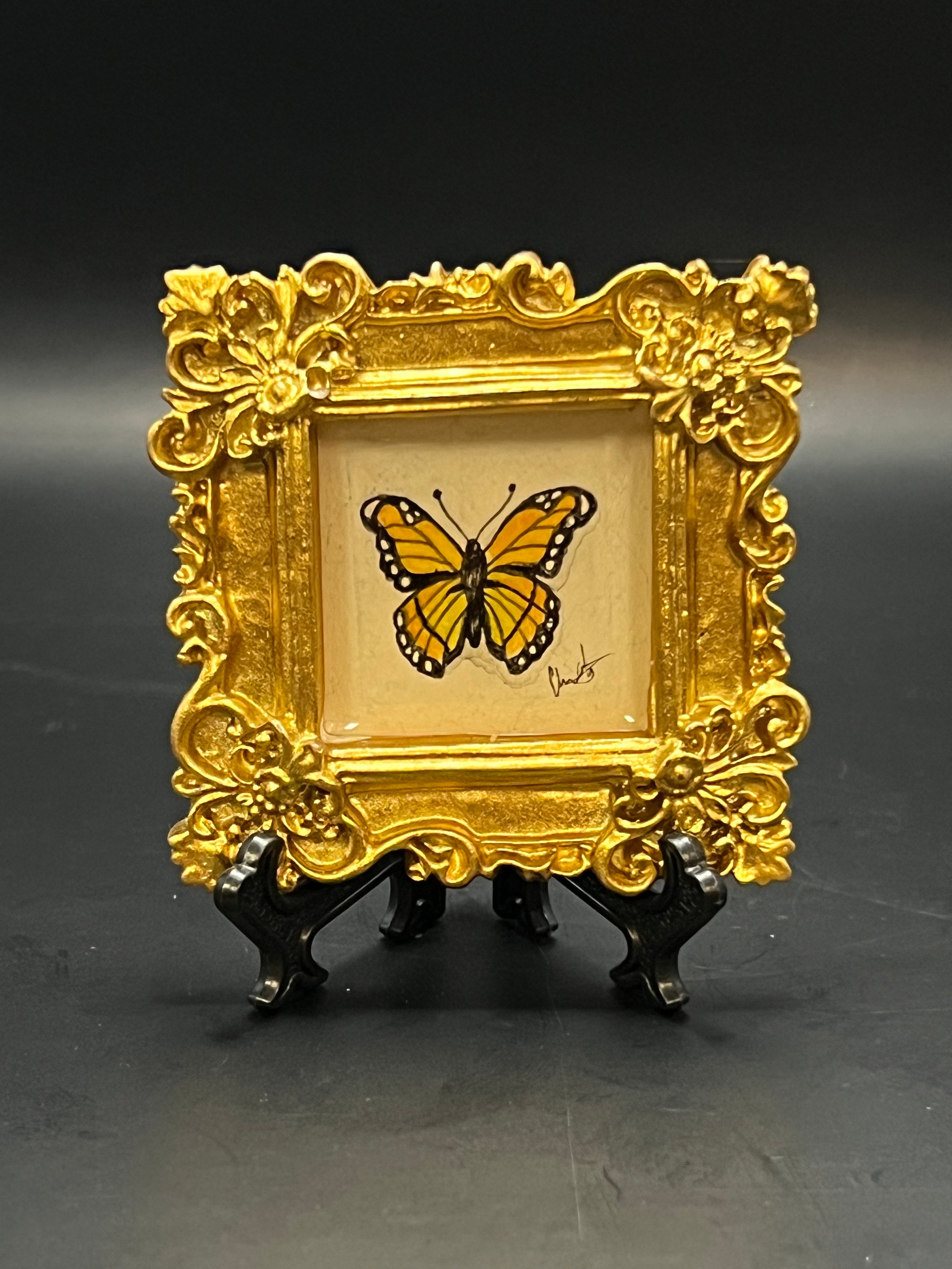 This is the Monarch Butterfly Mini Painting by Chris Foster Design. This showcases the front where a decorative gold frame with a yellow butterfly that includes black around it`s frame and white spots. The illustration is on a tan background with the studio's signature on a black stand. It is all against a dark background.