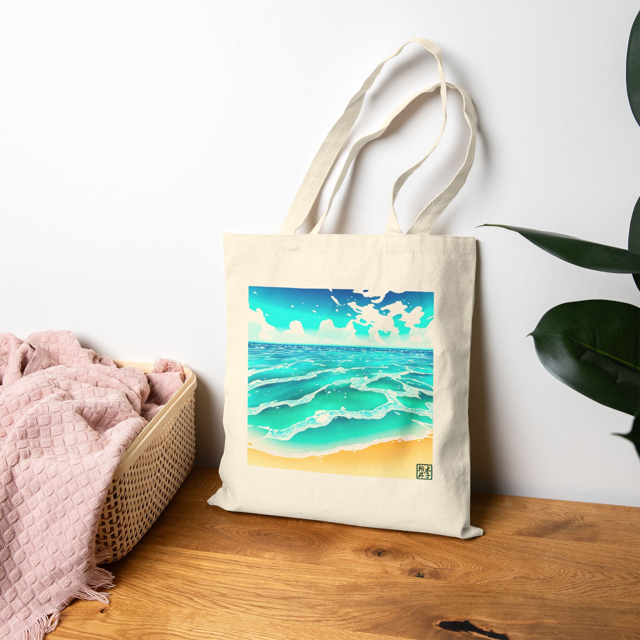 This is the Natural Beach Life Canvas Tote by Lee Hansheng Studios. The illustration is squared. The foreground has sand and the studio logo on the right corner outlined in blue. The waves are crashing into the sand with fluffly clouds in the back. There are mixes of blue, light blue, white, orange and tan colors. The tote is on a wooden surface with a pink blanket to the left and a plant to the right.