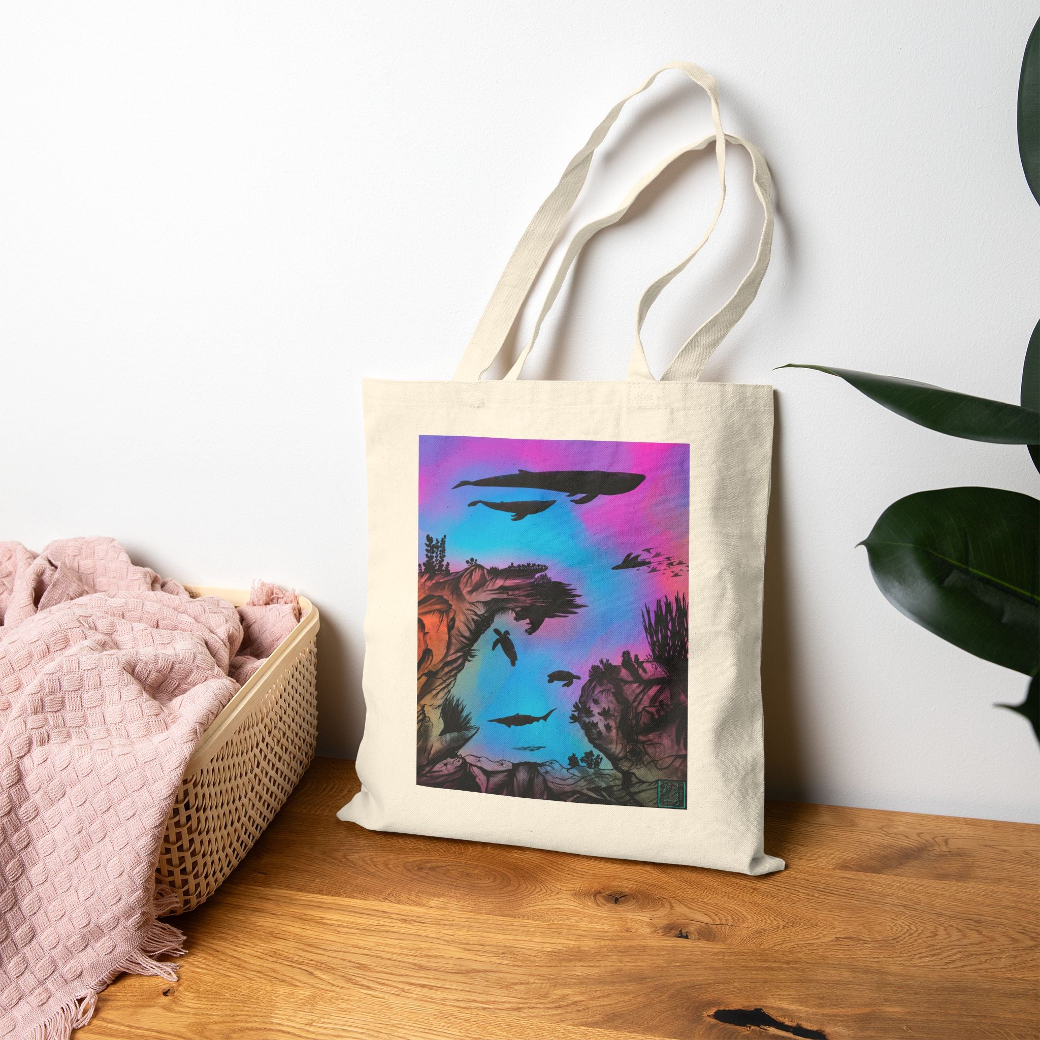 The Natural Into the Abyss Canvas Tote by Lee Hansheng Studios.The illustration is underwater seascape with aquatic plants on a marbled background.There are two blue whales,1 goblin shark,school of eagle rays,two sea turtles and a school of fish.On the bottom right corner where the front patch of the backpack there is the studio logo printed in black outlined in blue.The colors are pink,purple,blue and red/orange.The tote is on a wooden surface with a pink blanket to the left and plant to the right.