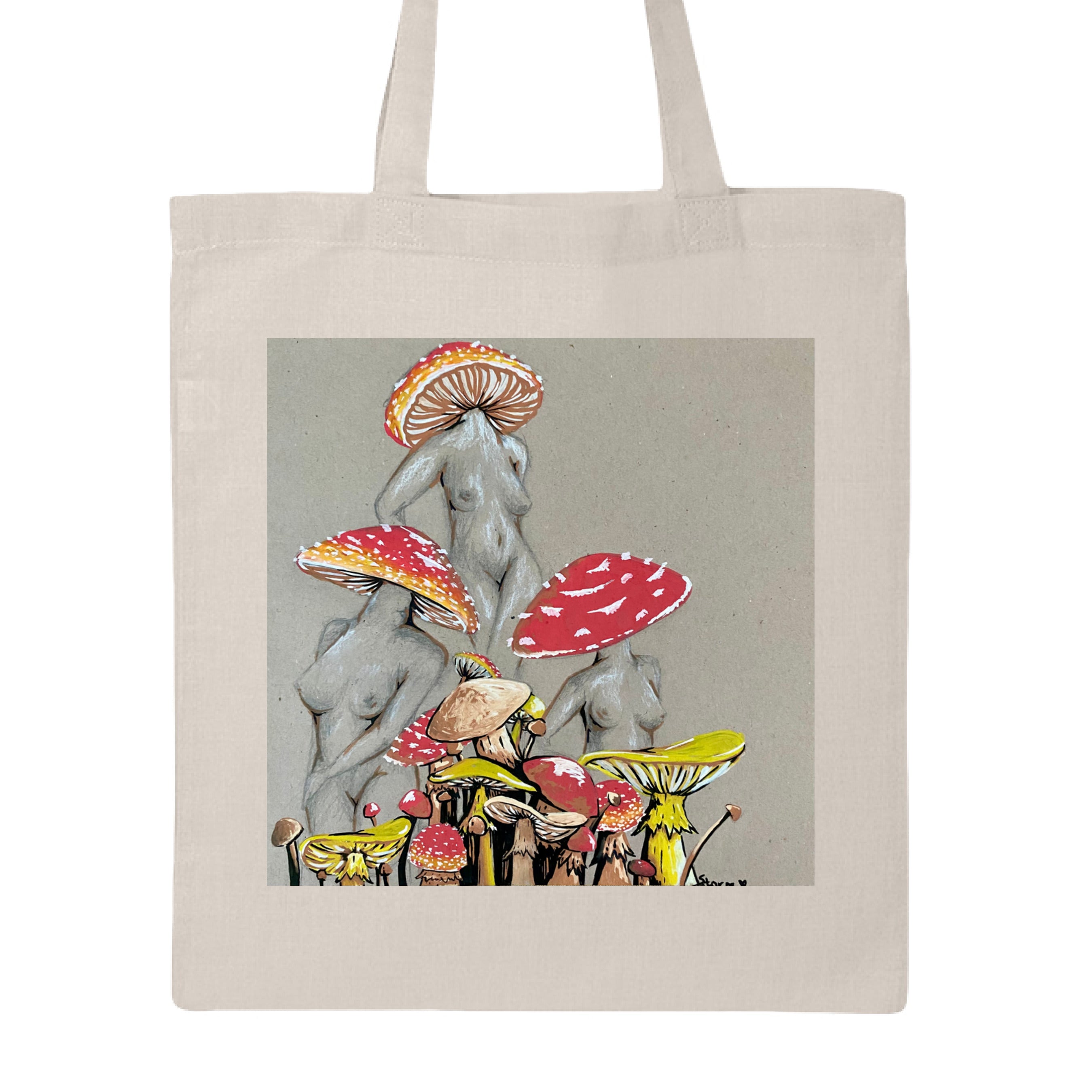 This is the Natural Miss Amanita Canvas Tote by Storm Garden Studio. This is showcasing the front. This illustration is on tan/grey background. There are three fusions of female human anatomy and mushroom fungi. Their heads are different styles of mushrooms, bodies are a gray/white outlined in brown. Below are different mushrooms and shapes with a mix of tan, red, white and yellow colors. The tote handles are color matched. It is against a white background.
