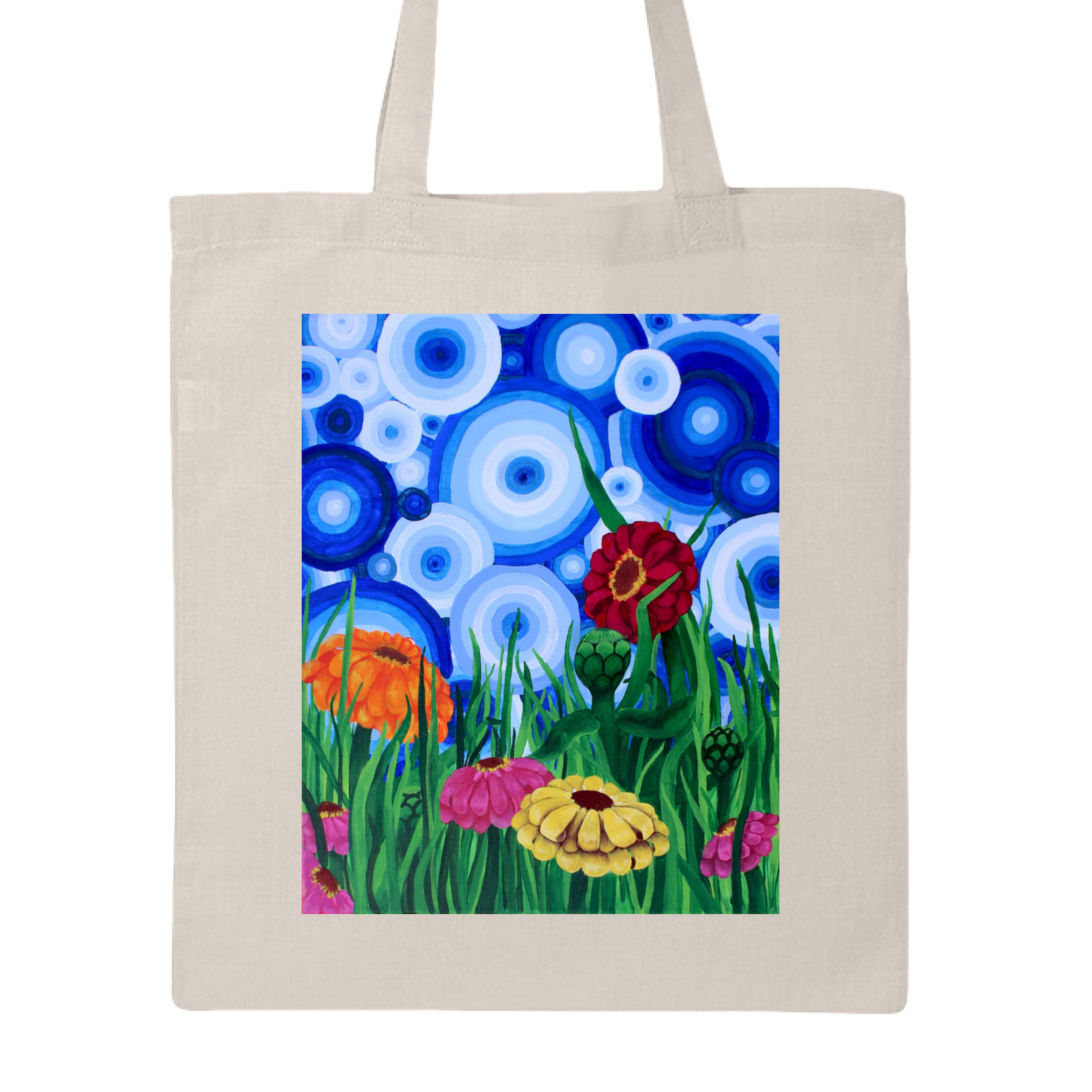 This is the Natural Zinnia 2 Canvas Tote by Storm Garden Studio. This showcases the front of the tote. The rectangular illustration are bold and color zinnias like red, yellow and orange across tall green grass. The background has many different circles in blue, dark blue and white. The tote is against a white background.
