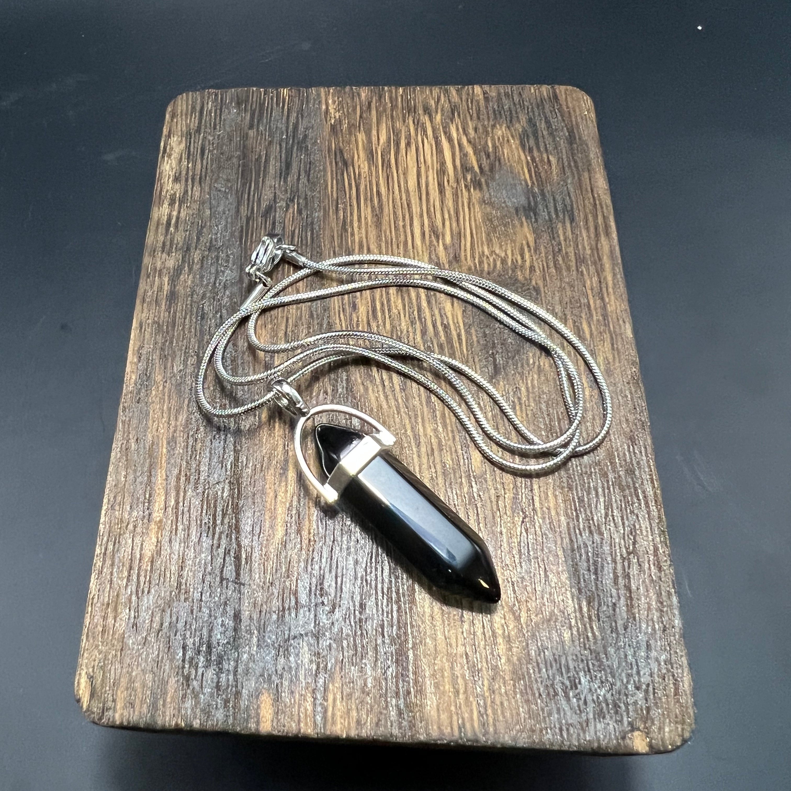 This is the Onyx Hexagon Stainless Steel Snake Chain Necklace by Lee Hansheng Studios. The necklace with a hexagon pendant is laying on a wooden surface against a dark background with the snake chain and clasp. 