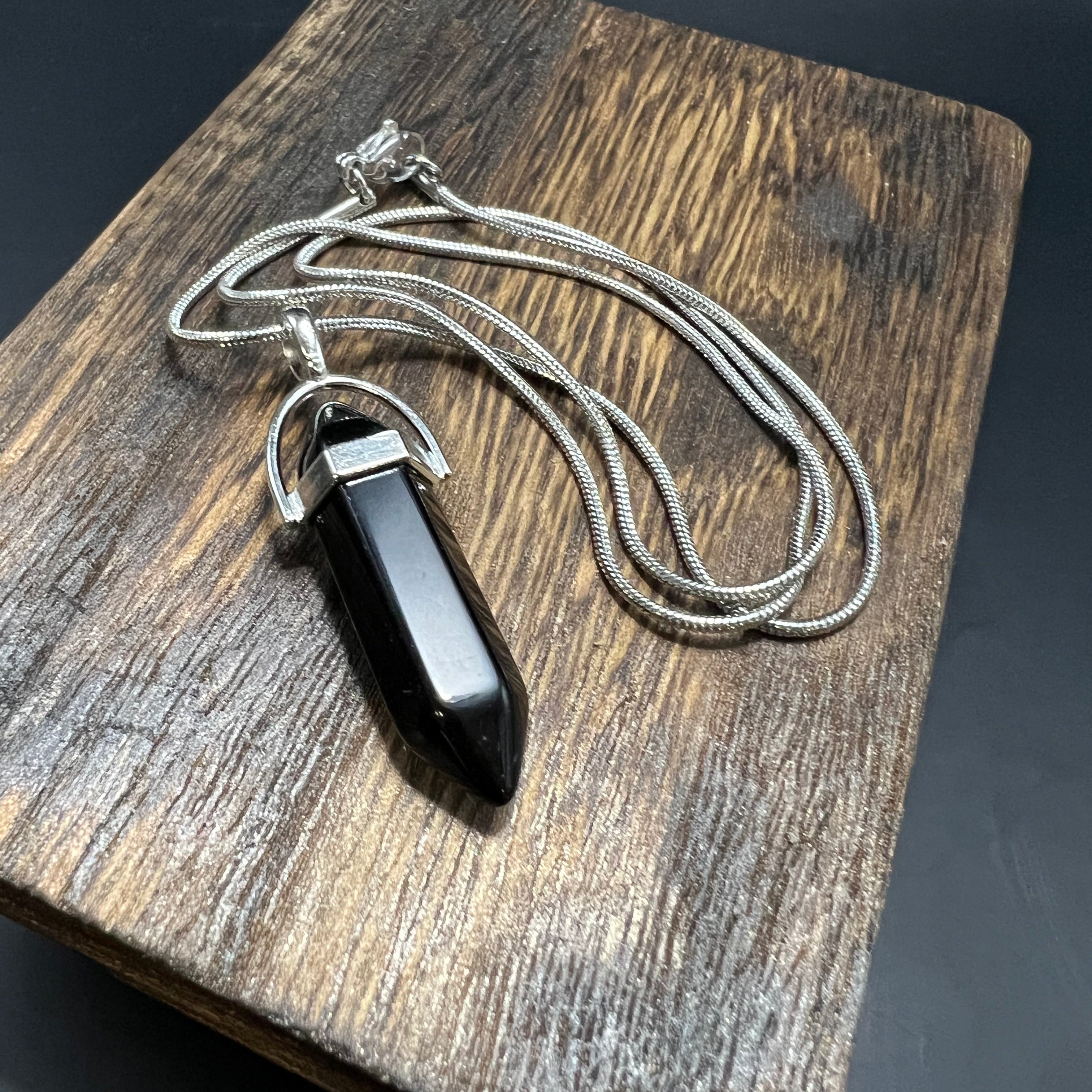 This is the Onyx Hexagon Stainless Steel Snake Chain Necklace by Lee Hansheng Studios. The necklace with a hexagon pendant is laying on a wooden surface against a dark background. 
