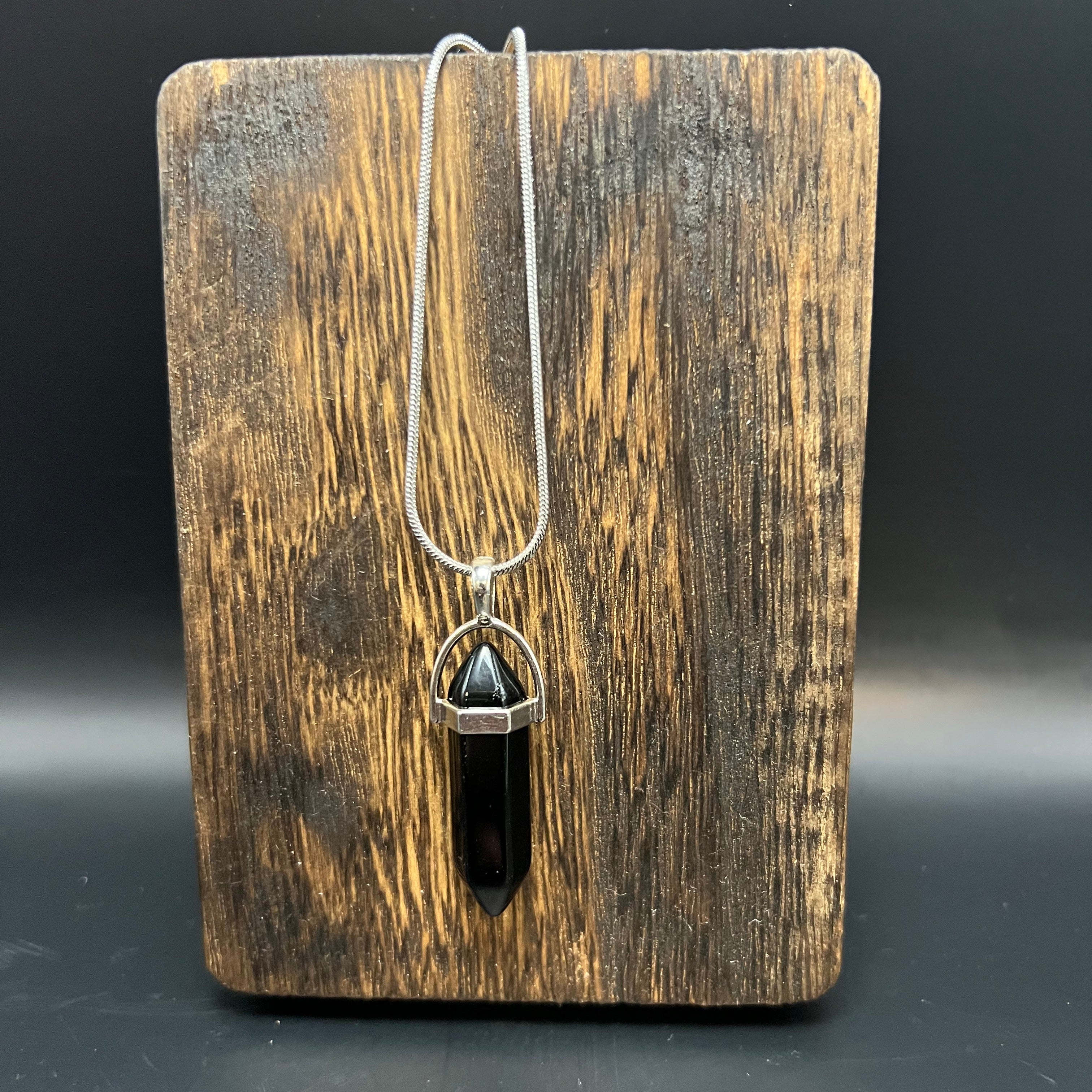This is the Onyx Hexagon Stainless Steel Snake Chain Necklace by Lee Hansheng Studios. The necklace with a hexagon pendant is hanging and against a wooden surface against a dark background. 