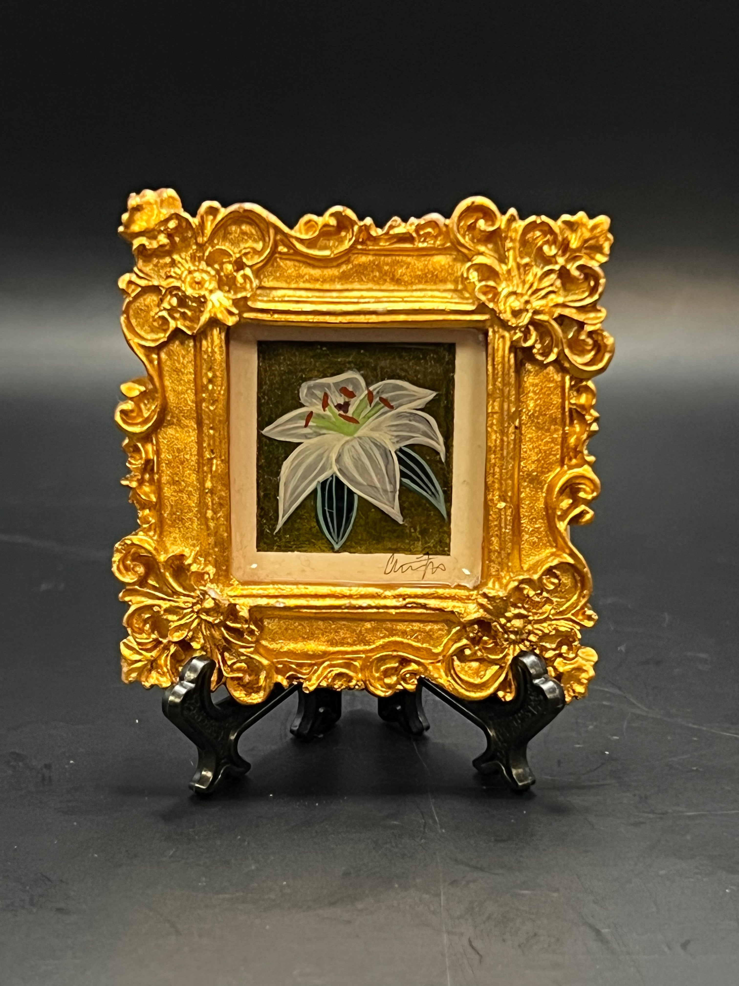 This is the Oriental Lily Mini Painting by Chris Foster Design. This is showcasing the front of the painting. This is a decorative gold frame with a small painting of a white flower, two dark green leaves on a green/grey background on a black stand. It is against a dark background.